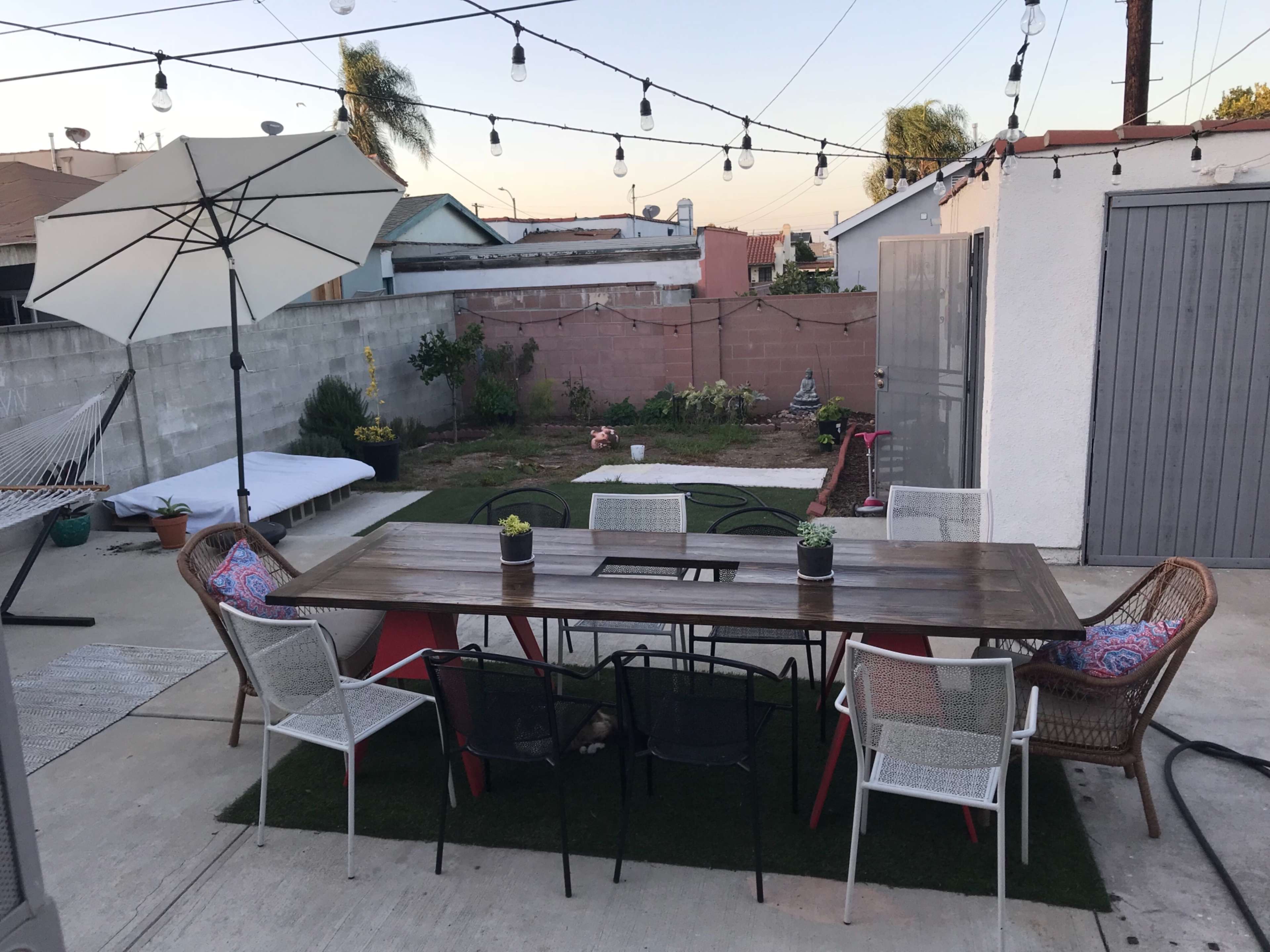 A backyard patio features a large wooden table surrounded by various chairs, an umbrella for shade, and garden beds with plants in the background.