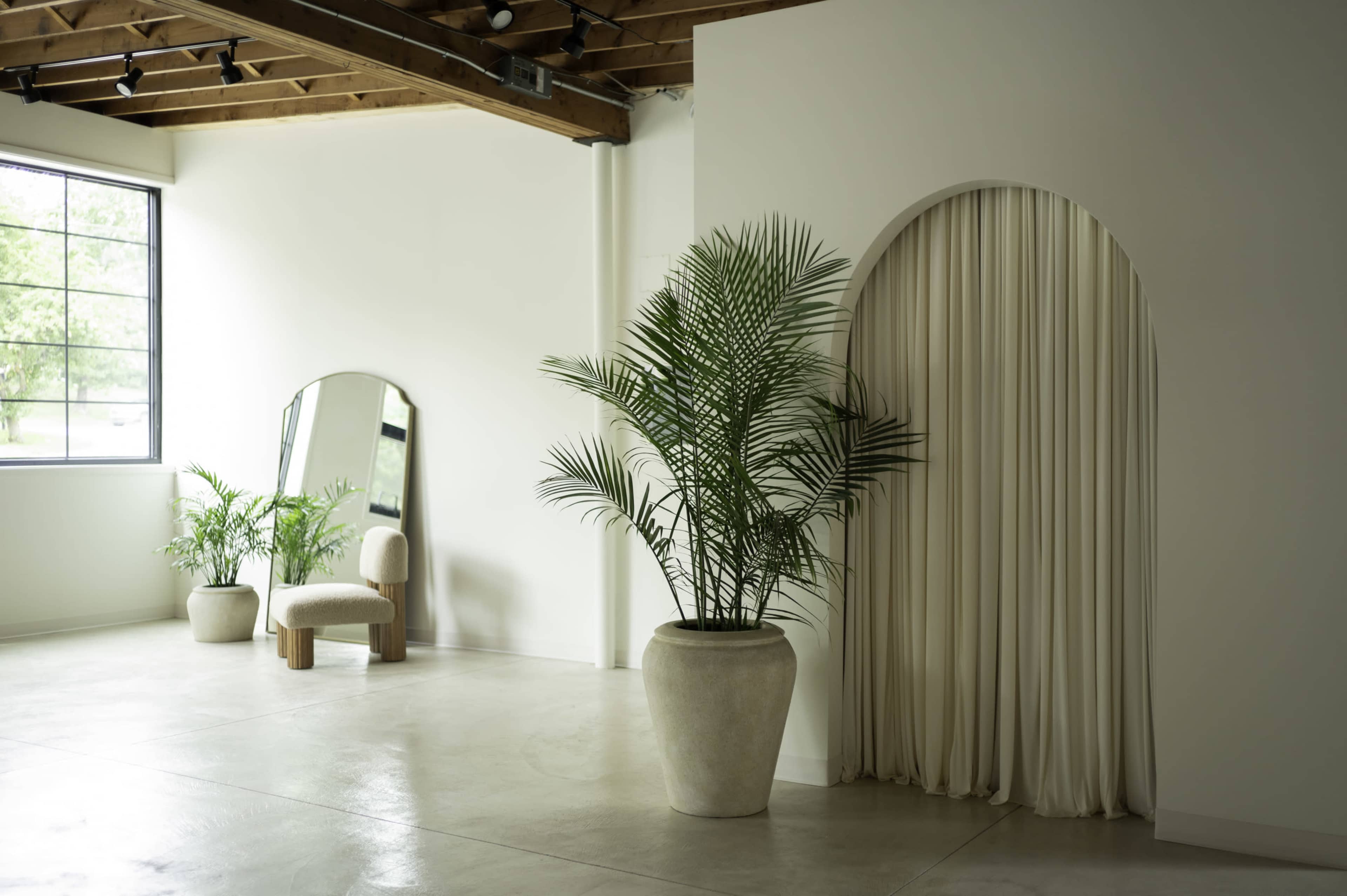 The image shows a minimalistic interior space featuring a large window, a mirror, a potted plant, and a curtain draped in an arched wall opening.
