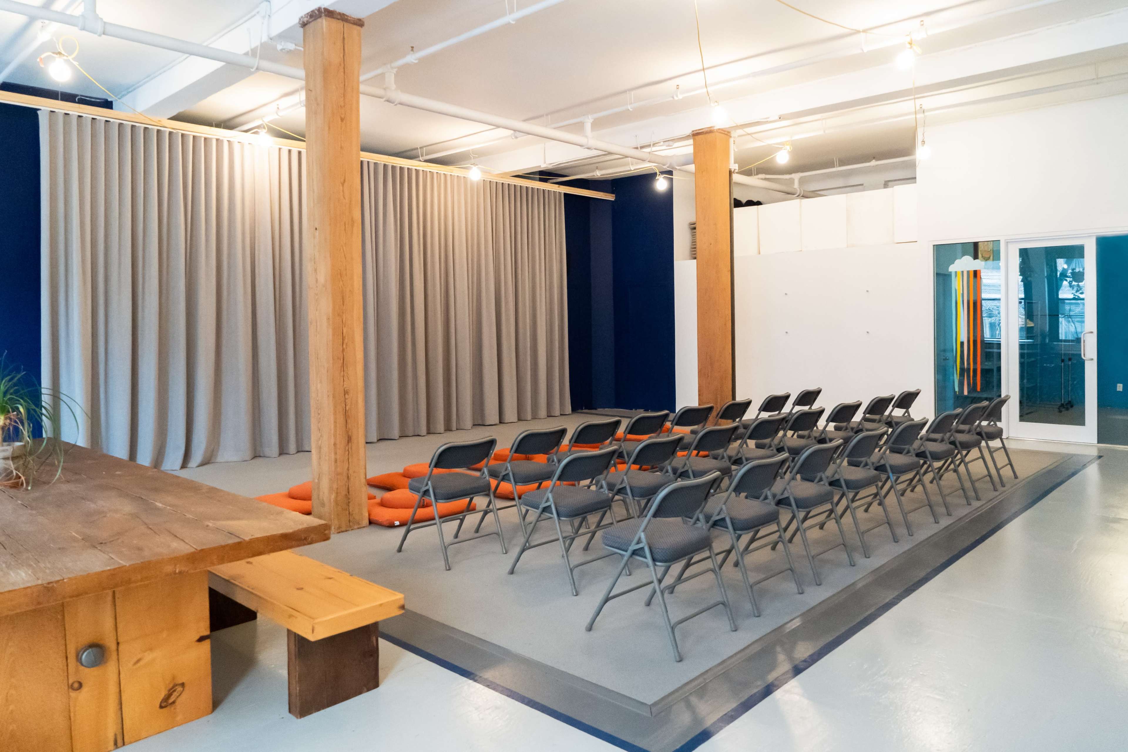 A spacious room set up for an event, featuring rows of folding chairs facing a curtain-draped stage area, with a wooden table to the side.