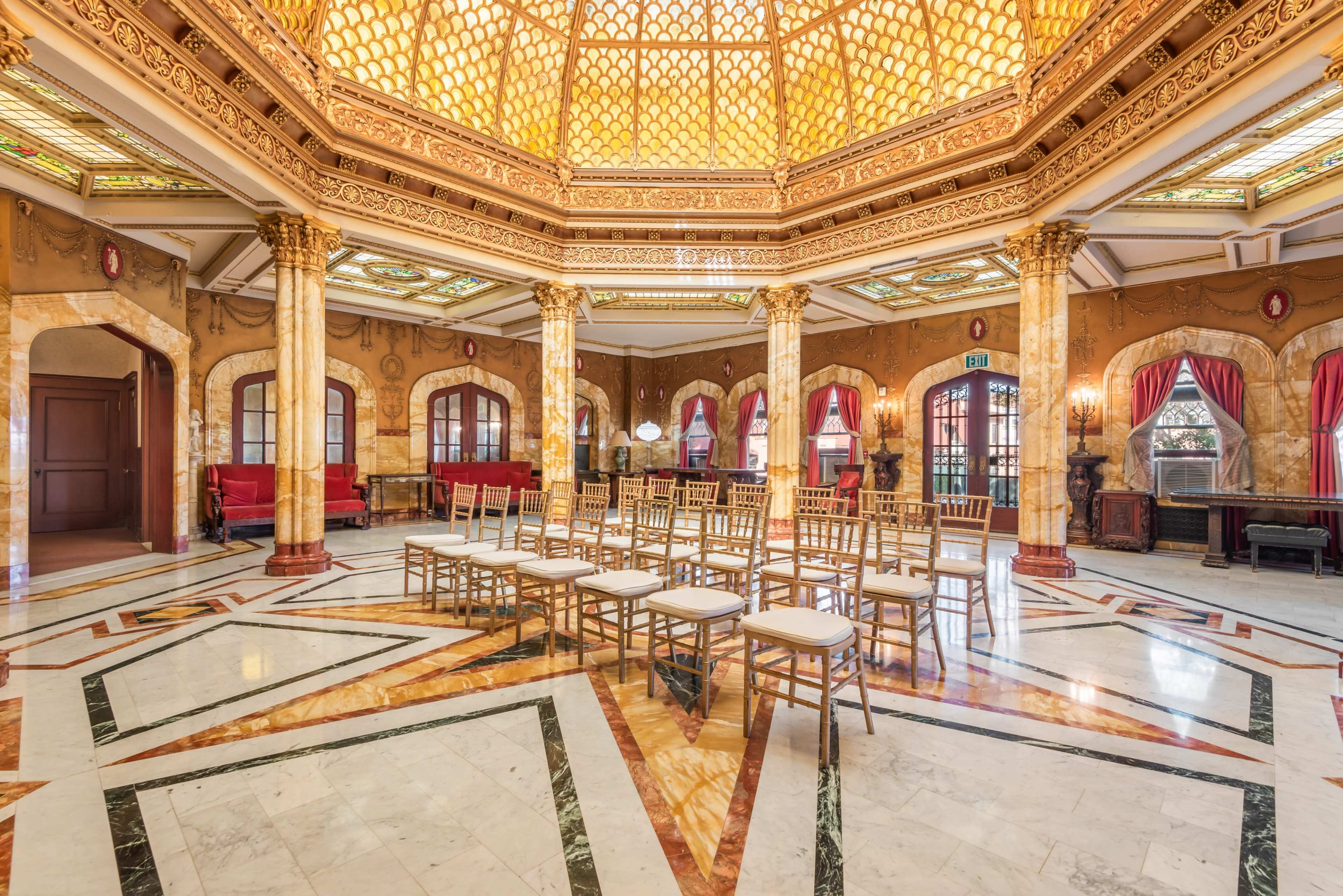 The image shows an ornate, well-lit hall with a patterned marble floor, large windows, and rows of empty chairs arranged in the center.