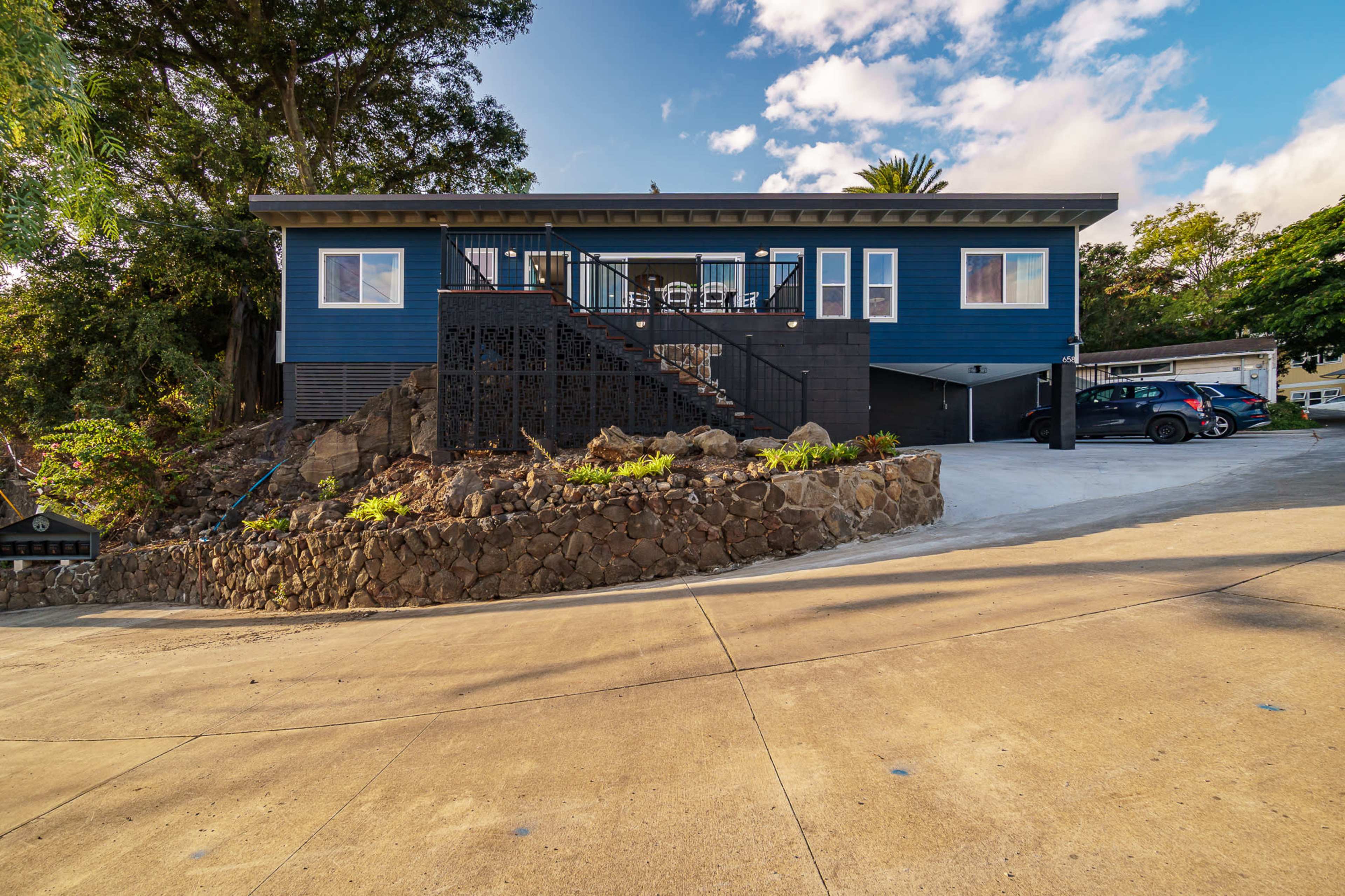 A blue house sits on a sloped driveway surrounded by rocks and greenery, with a few vehicles parked below.