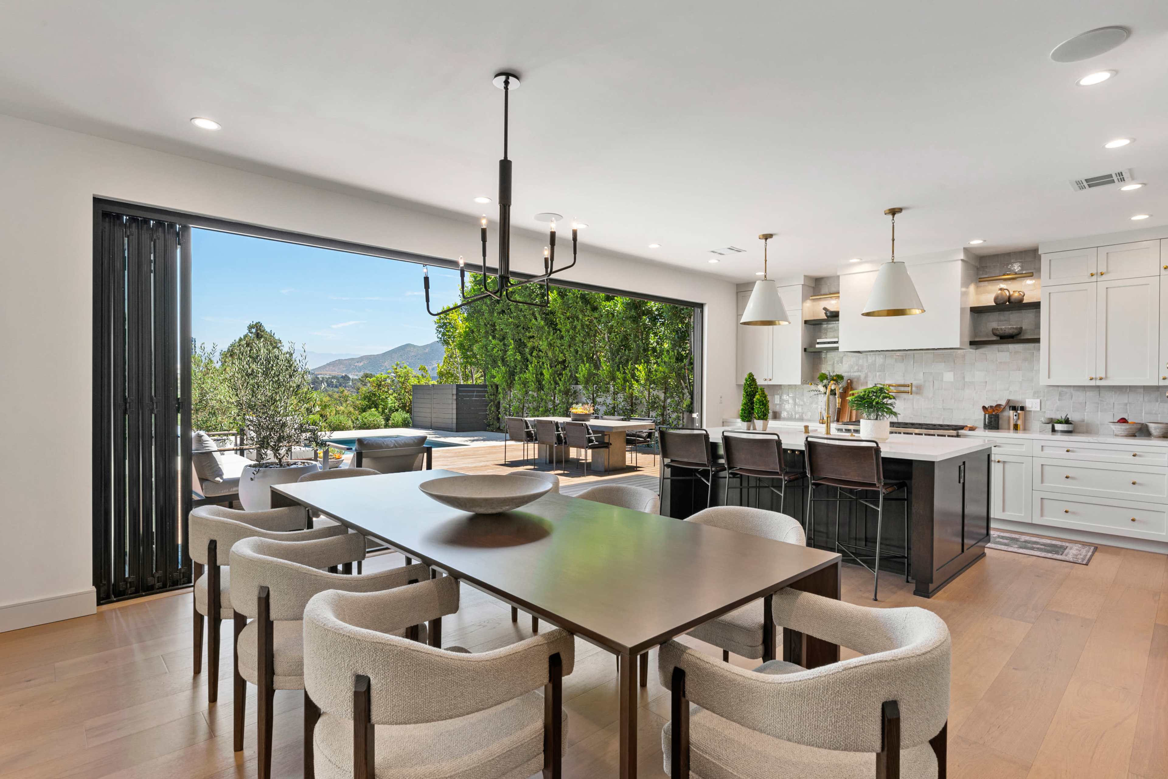 A modern kitchen and dining area features a large table surrounded by upholstered chairs, with an open wall showcasing a view of the outdoor space and mountains in the background.