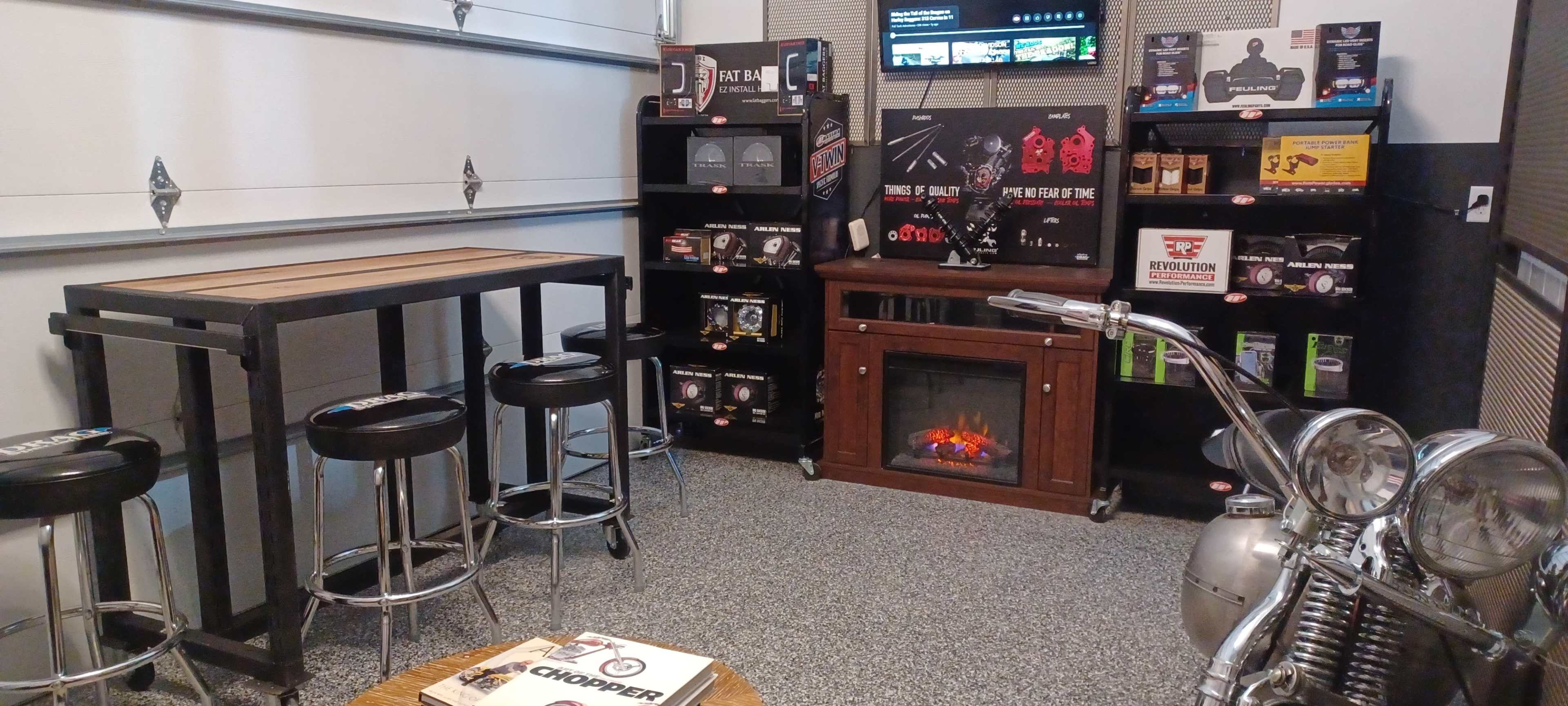 A garage setup featuring a wooden table, metal bar stools, a decorative electric fireplace, a display shelf with various products, and a motorcycle on the side.