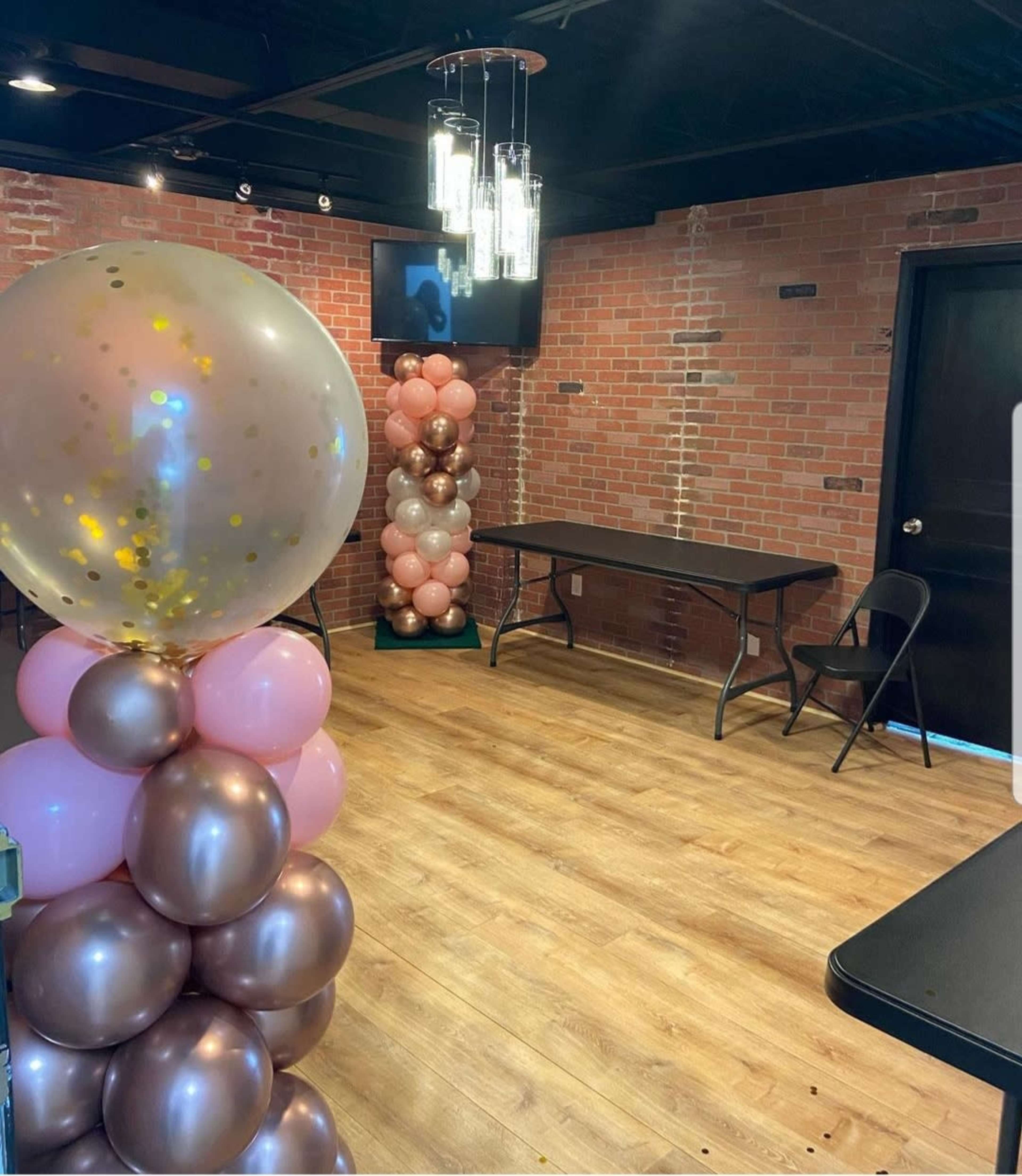The room features two tall balloon columns in pink and brown, a television on the wall, and two black folding tables with chairs.
