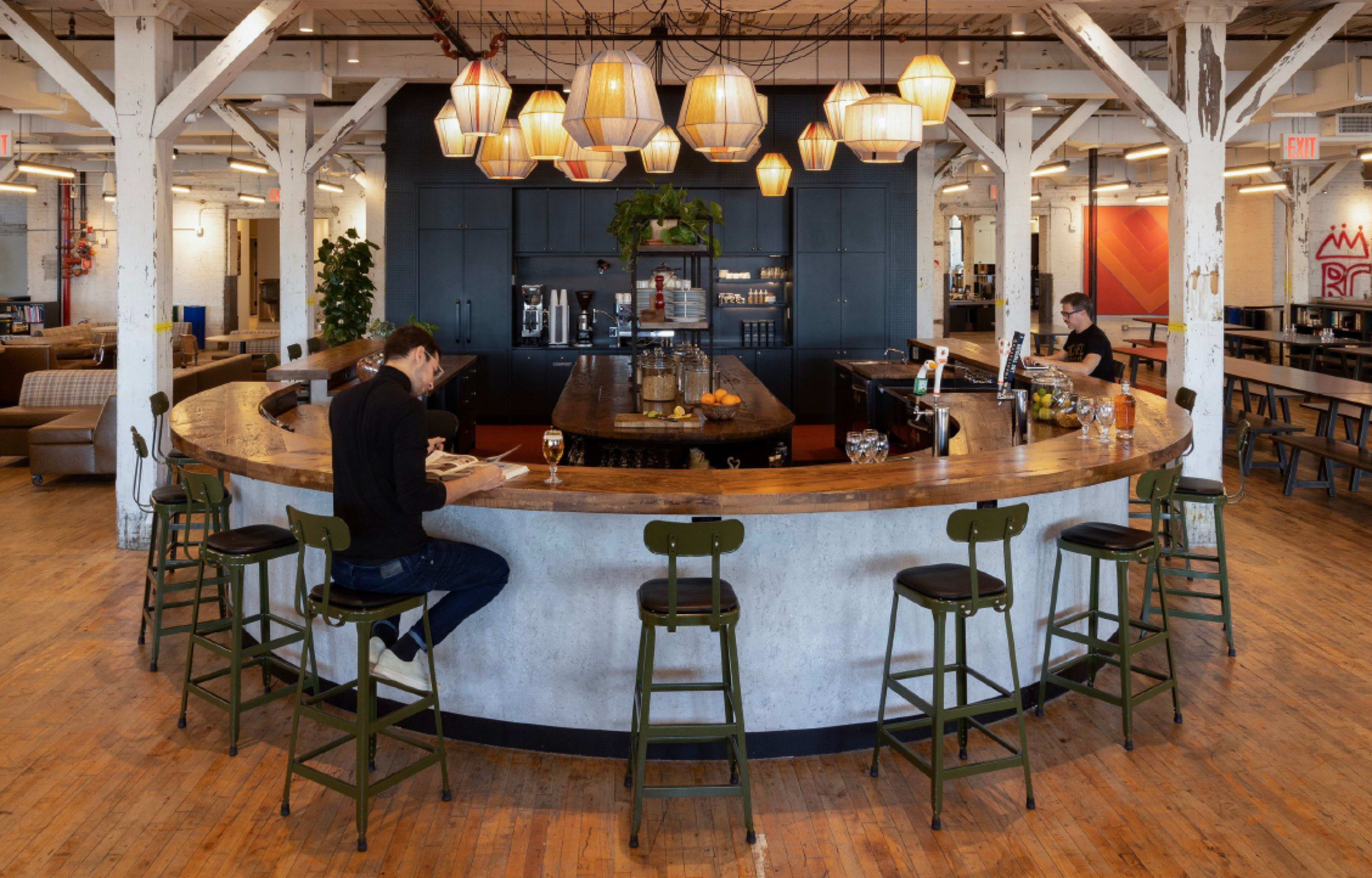 A modern café features a curved wooden bar with several patrons seated on tall stools, surrounded by warm pendant lighting and a sleek black background.
