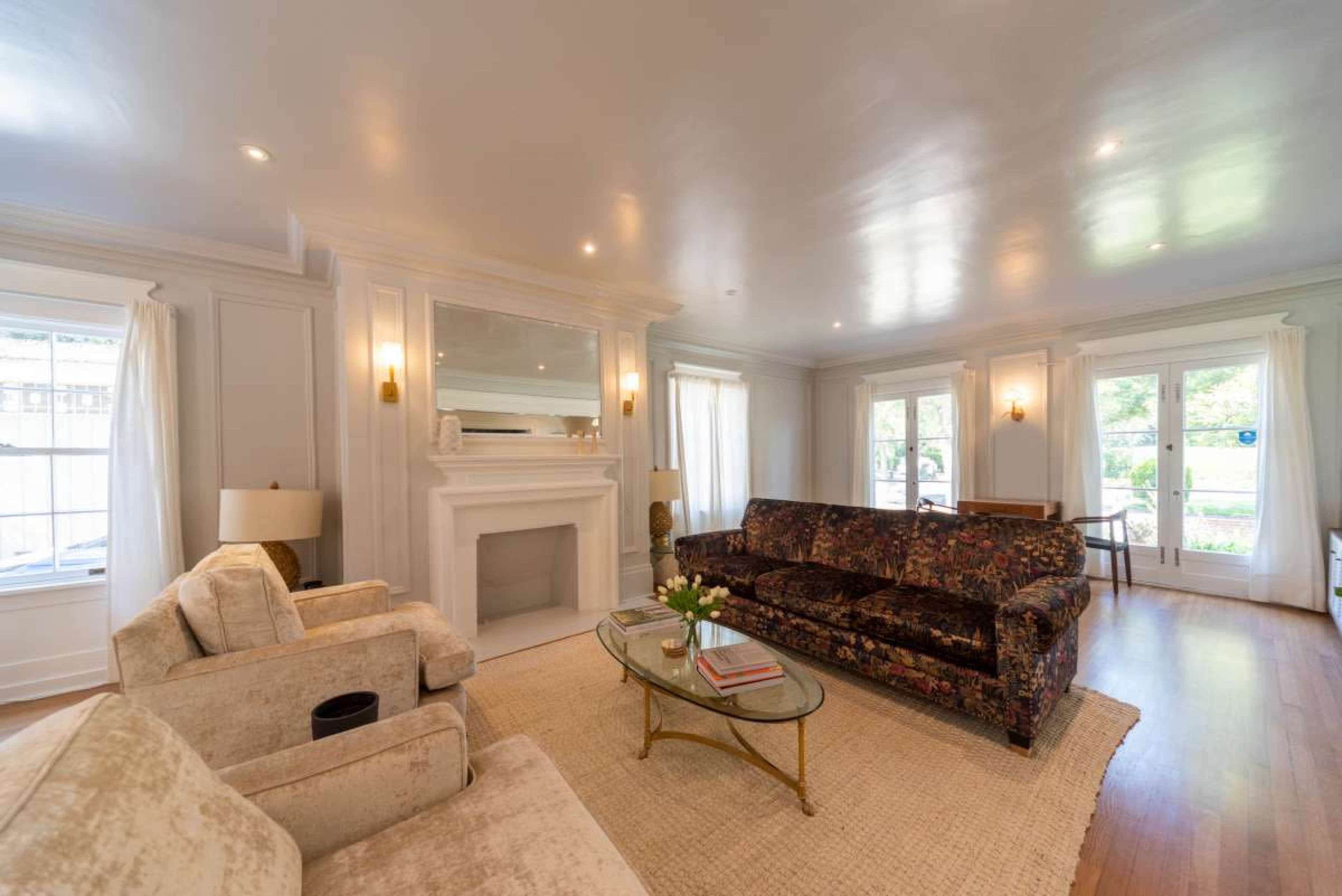 The image shows a brightly lit living room with a floral-patterned sofa, a glass coffee table, and large windows allowing natural light.