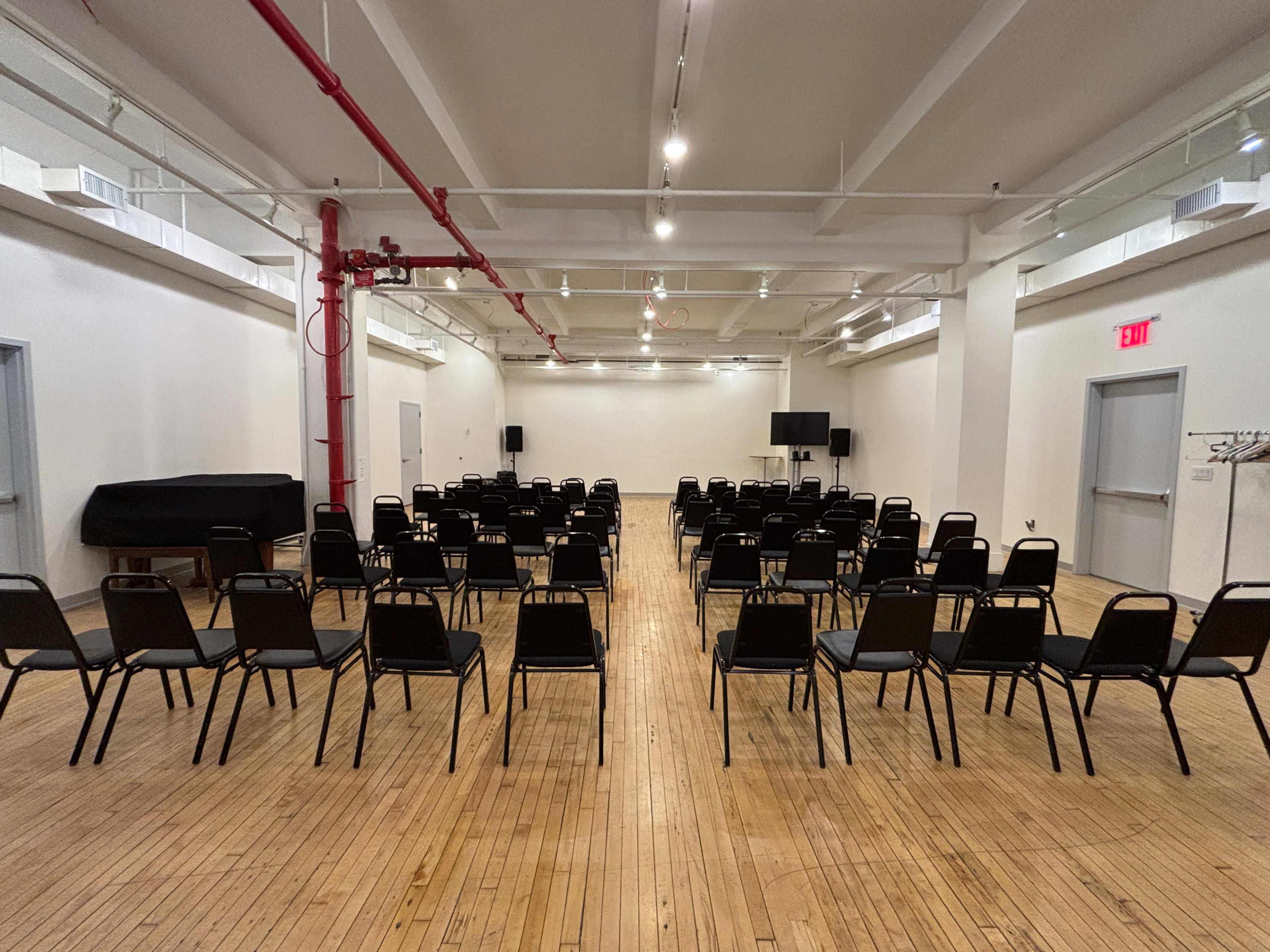 The image shows a large, empty room arranged with rows of black chairs facing a stage area, with a wooden floor and gray walls.