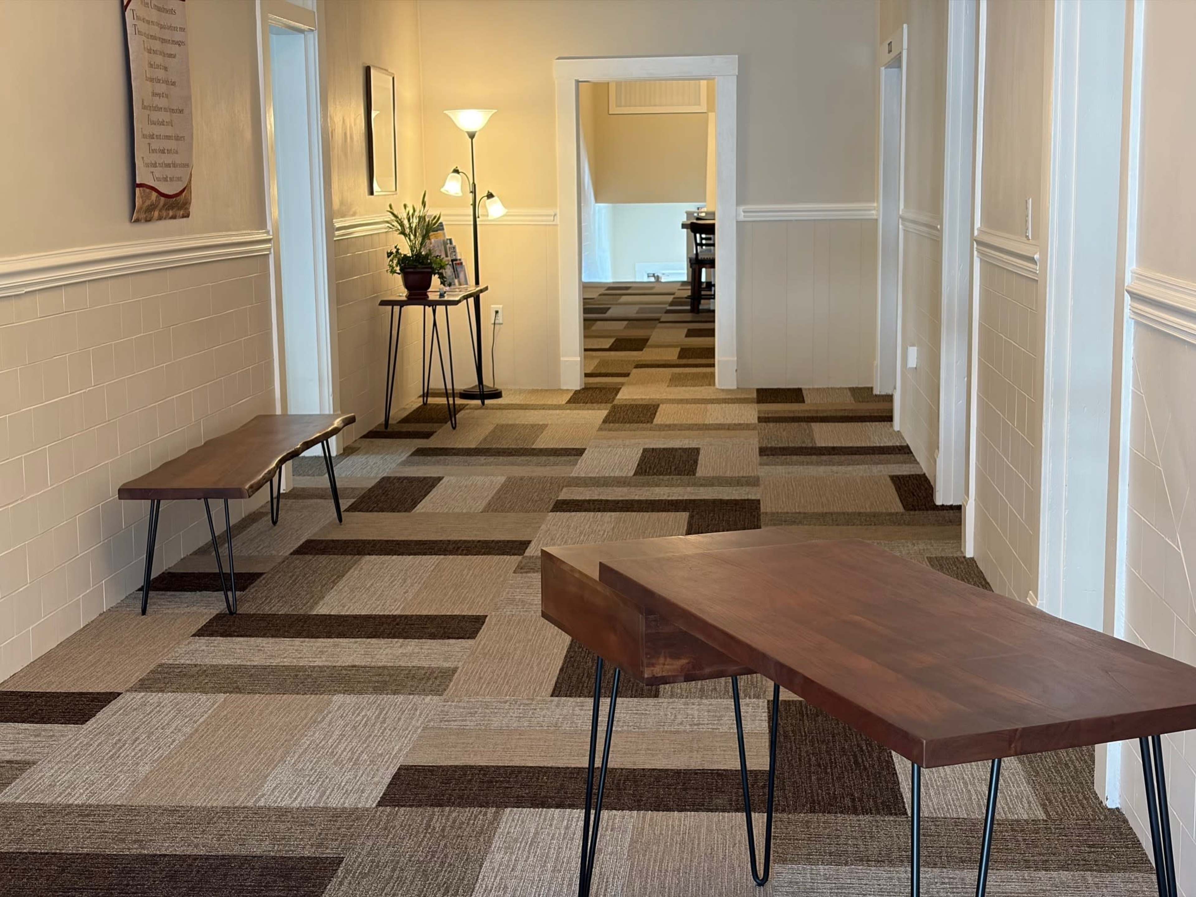 The image shows a hallway with light-colored walls and a patterned carpet, featuring two wooden tables with thin legs and a small potted plant on one table.