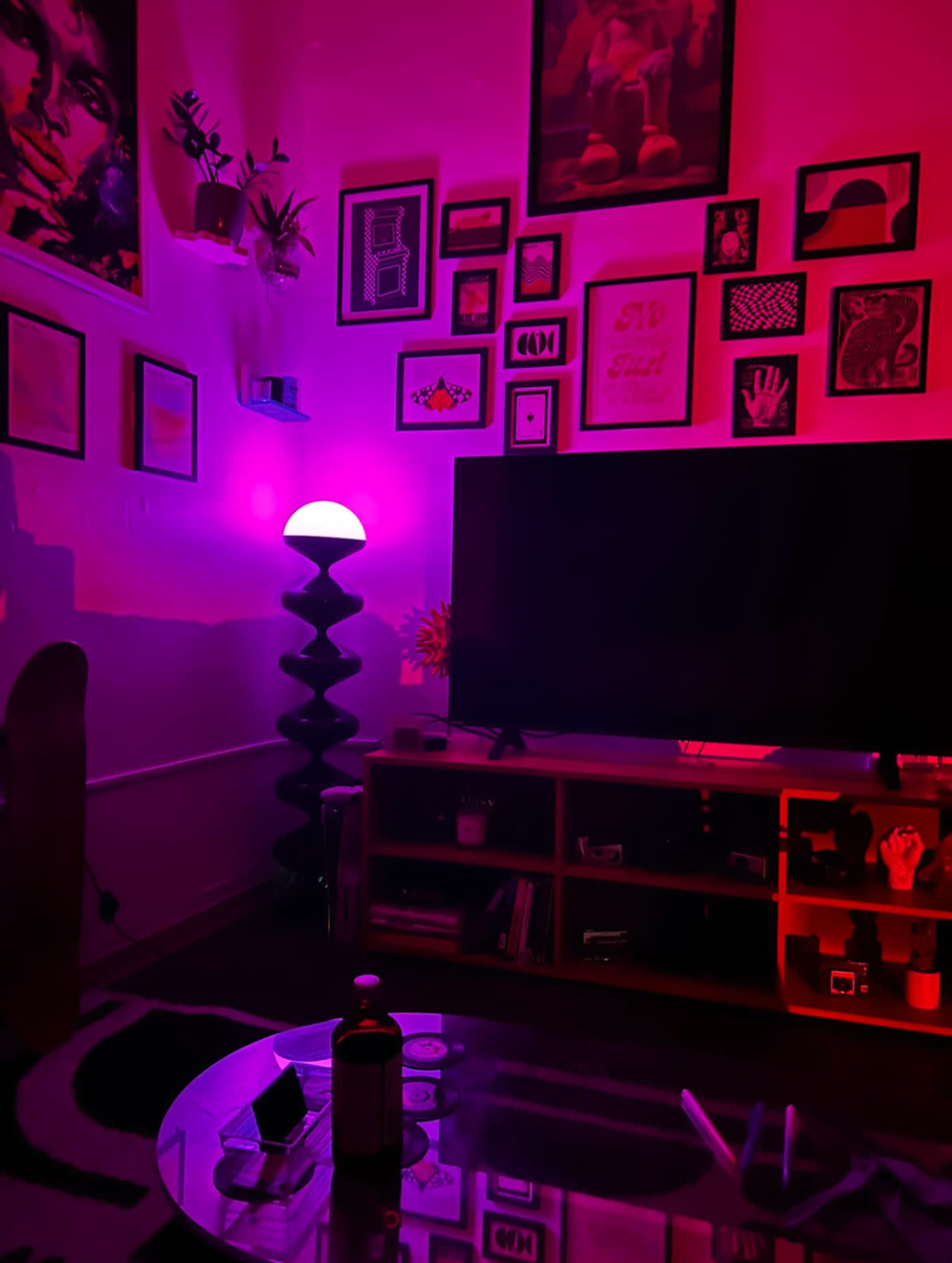 The image shows a dimly lit room featuring a large television, a pink lamp, colorful wall art, and a coffee table with various items.
