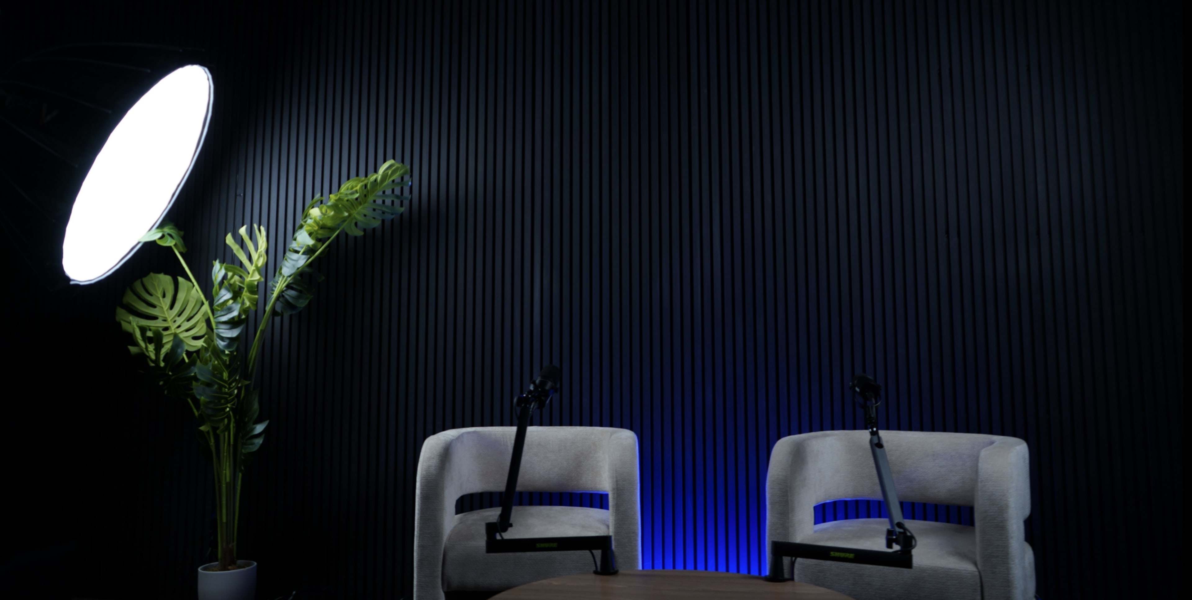 The image shows a minimalist indoor setting with two empty chairs, a round table, a large light source, and a potted plant against a textured black wall.