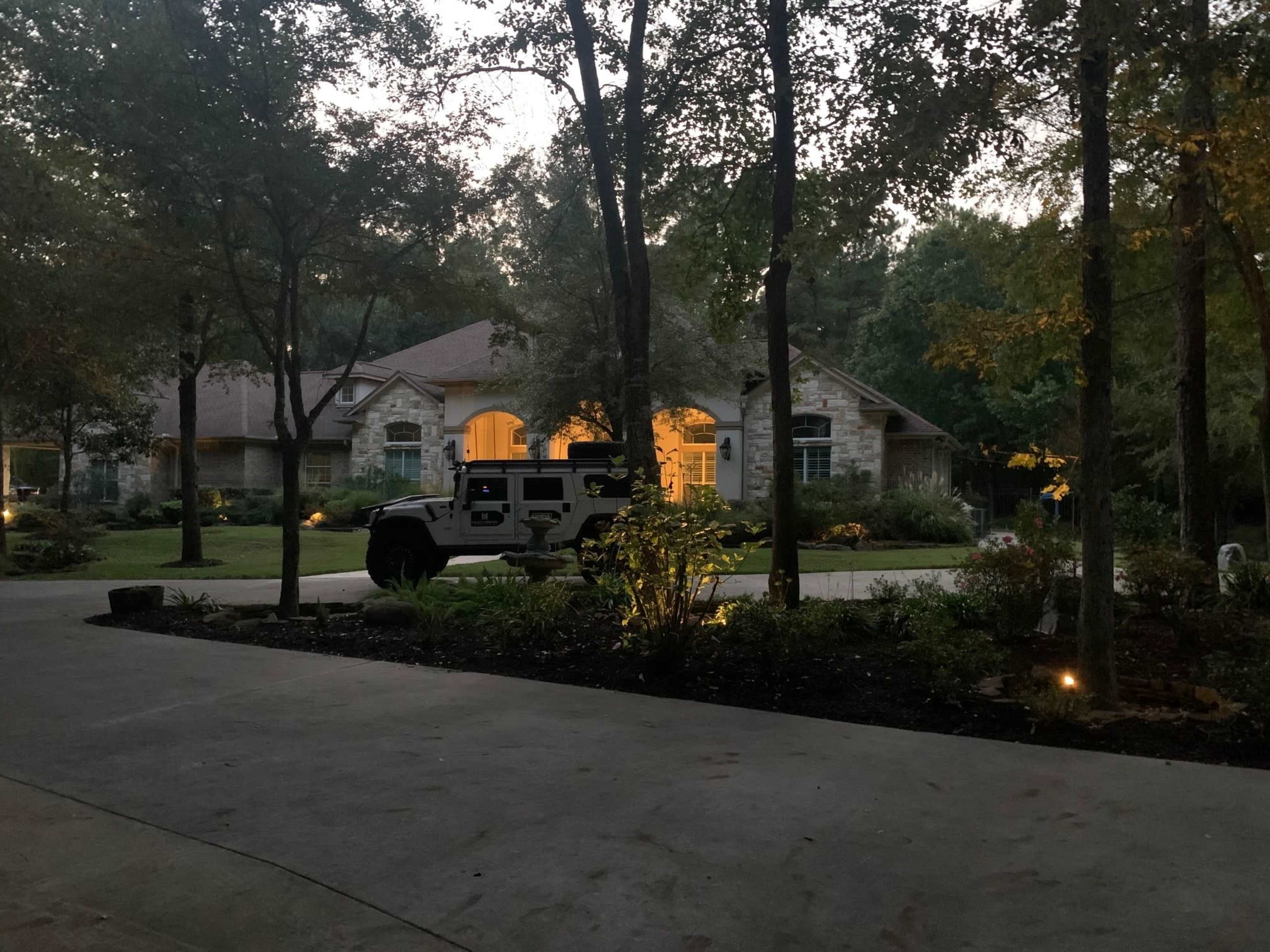 A suburban house with a driveway lined by trees is illuminated by porch lights as a vehicle is parked in front.