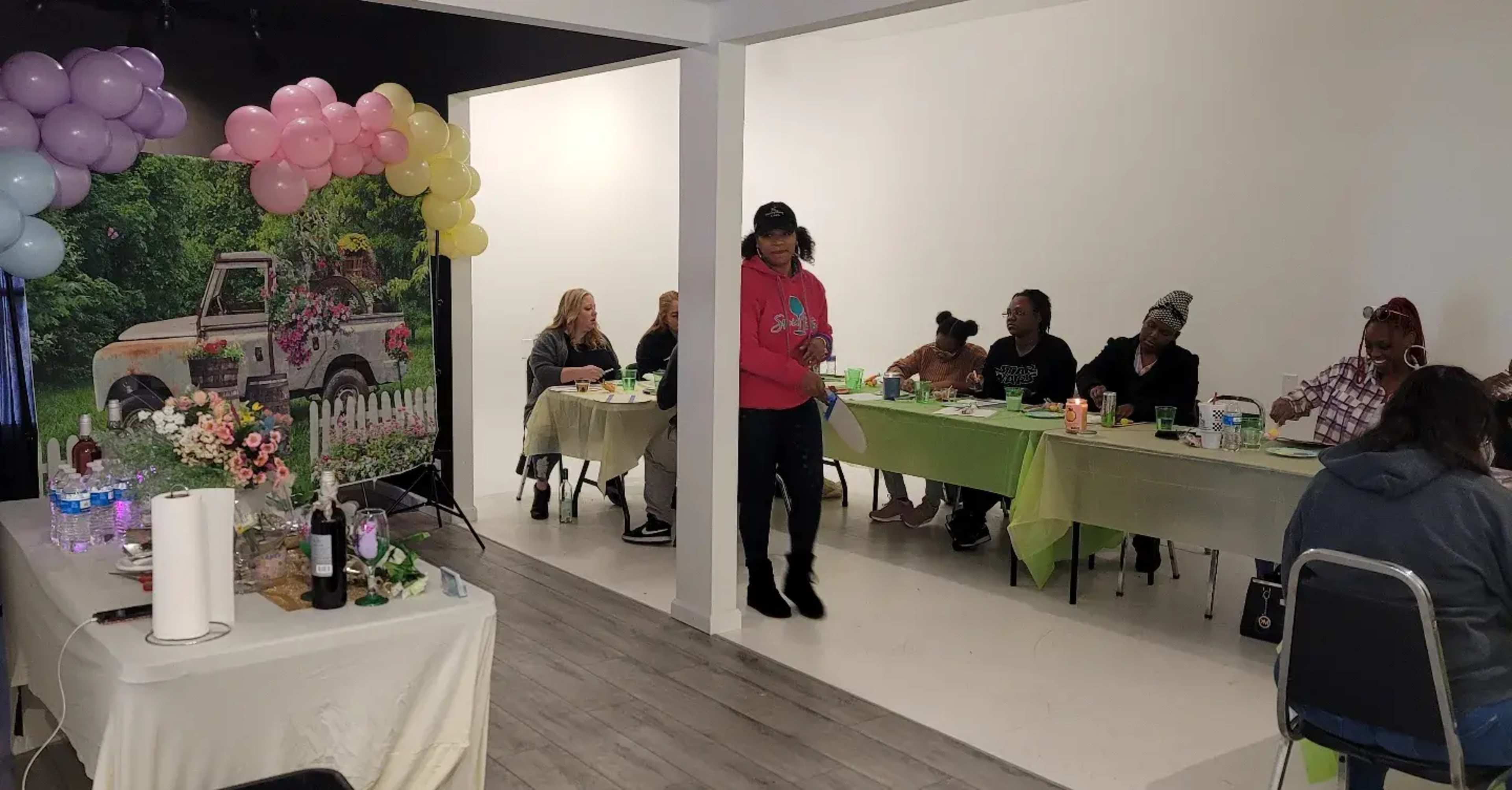 A group of people sits at tables in a room decorated with a floral backdrop and balloons, while one individual stands and interacts with others.