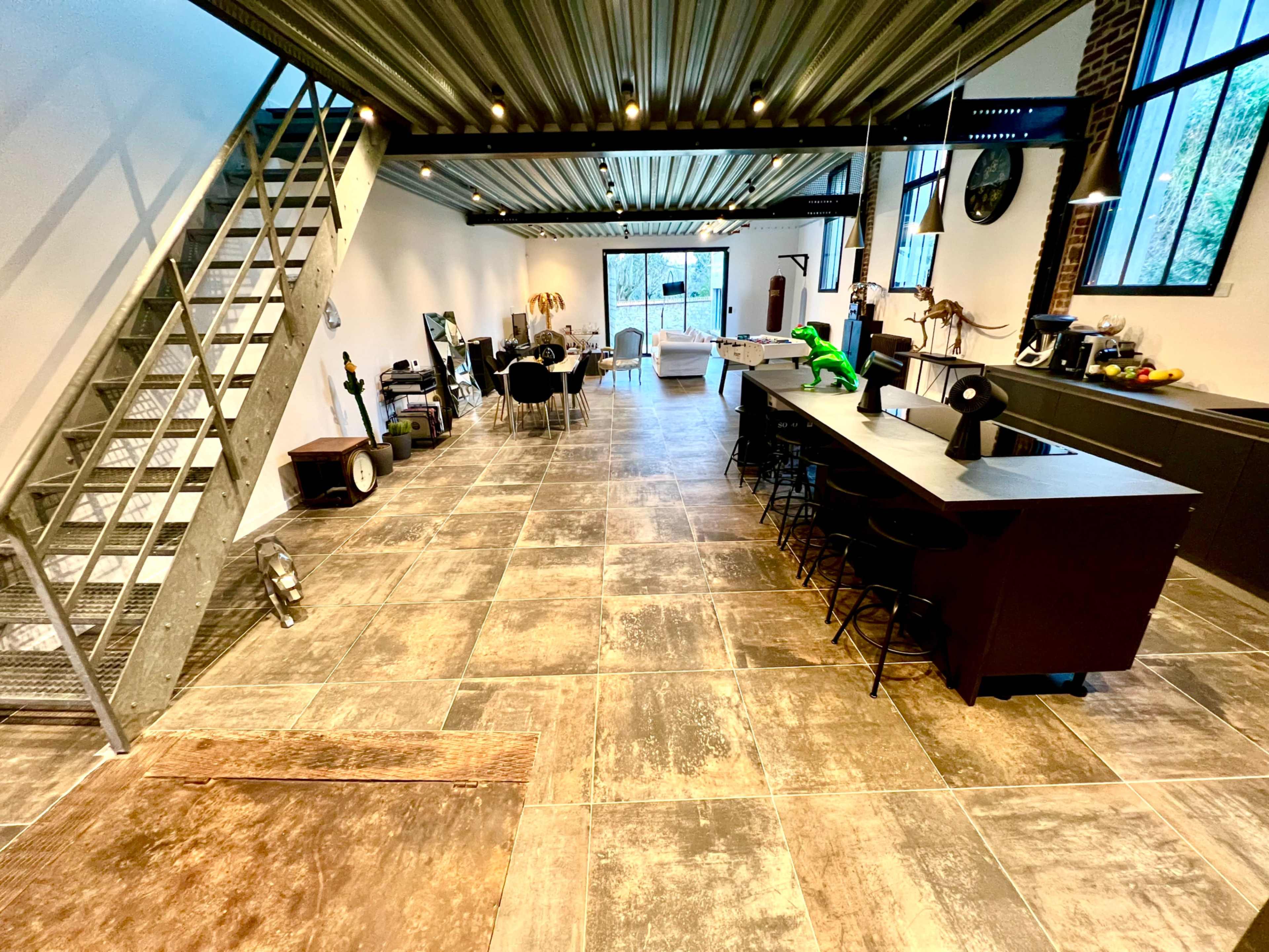 The image shows a spacious, modern interior of a warehouse-style office, featuring an open layout with a staircase, dining area, and kitchen bar along one side.