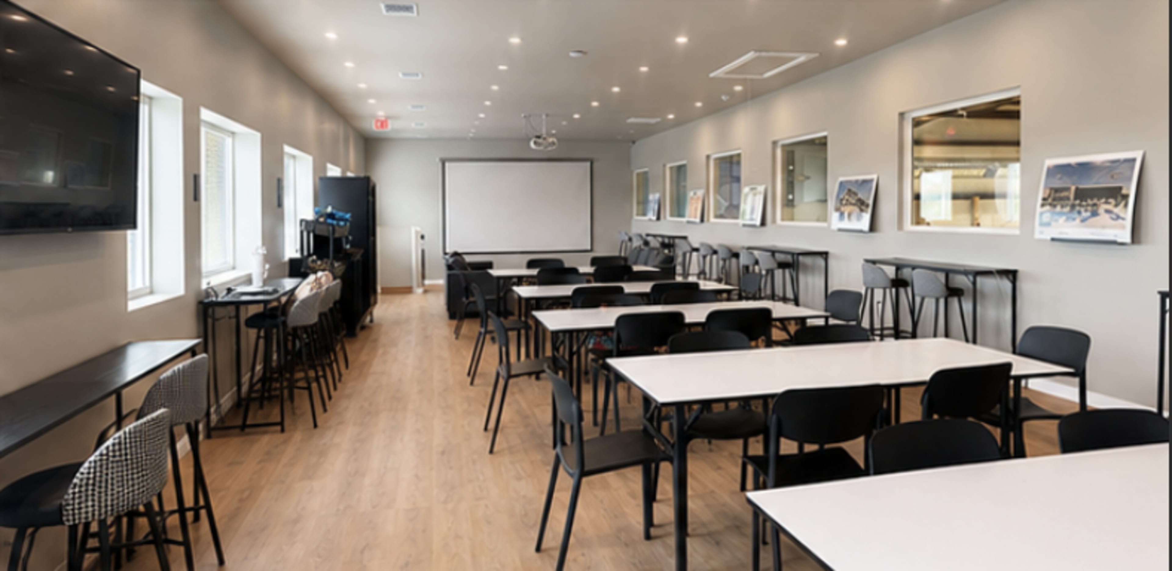 The image shows a modern classroom or meeting room set up with several white tables, black chairs, and a large screen at the front.