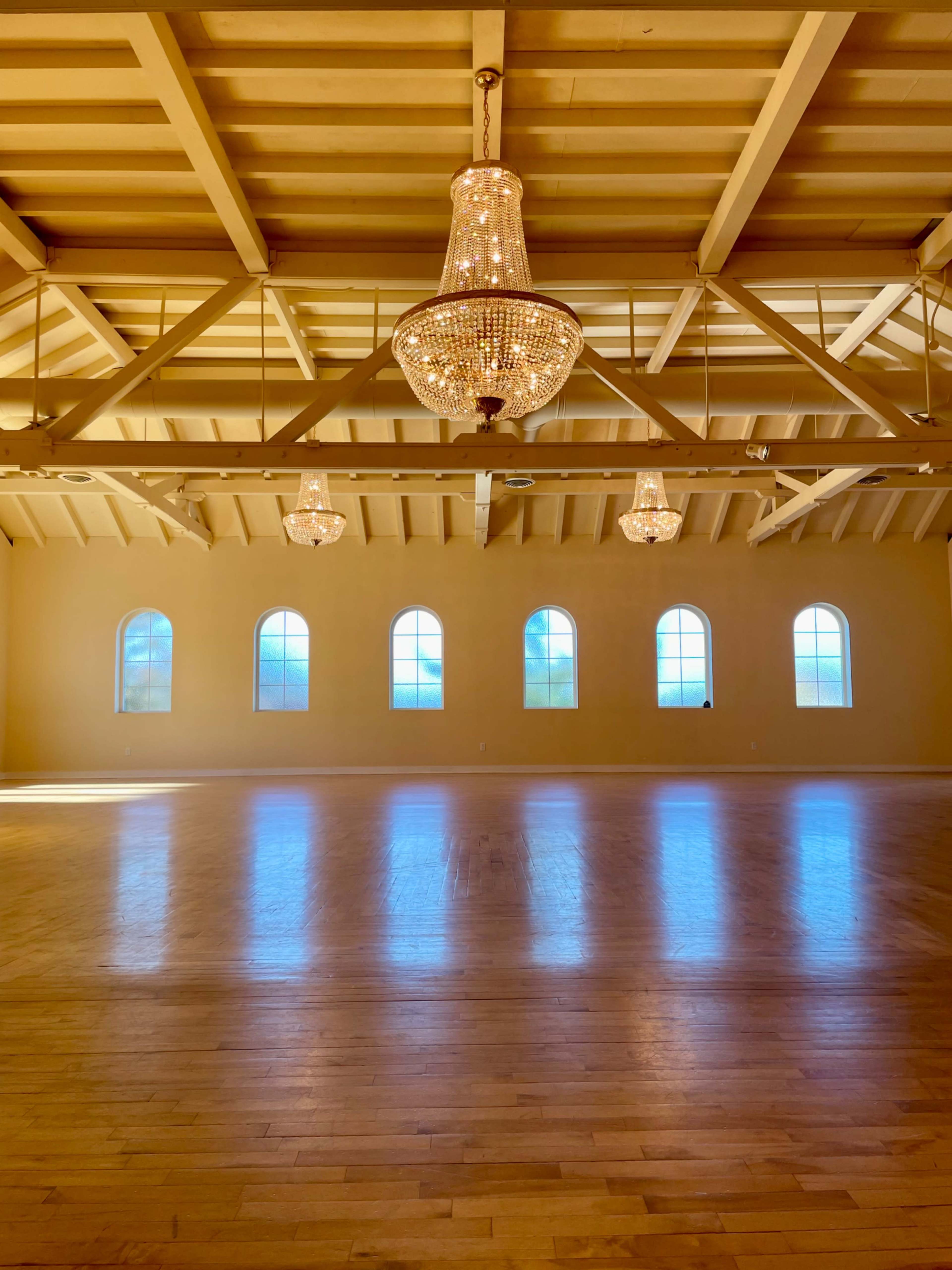 The image shows a spacious wooden room with a polished floor, featuring three large chandeliers and several arched windows allowing natural light to flood in.