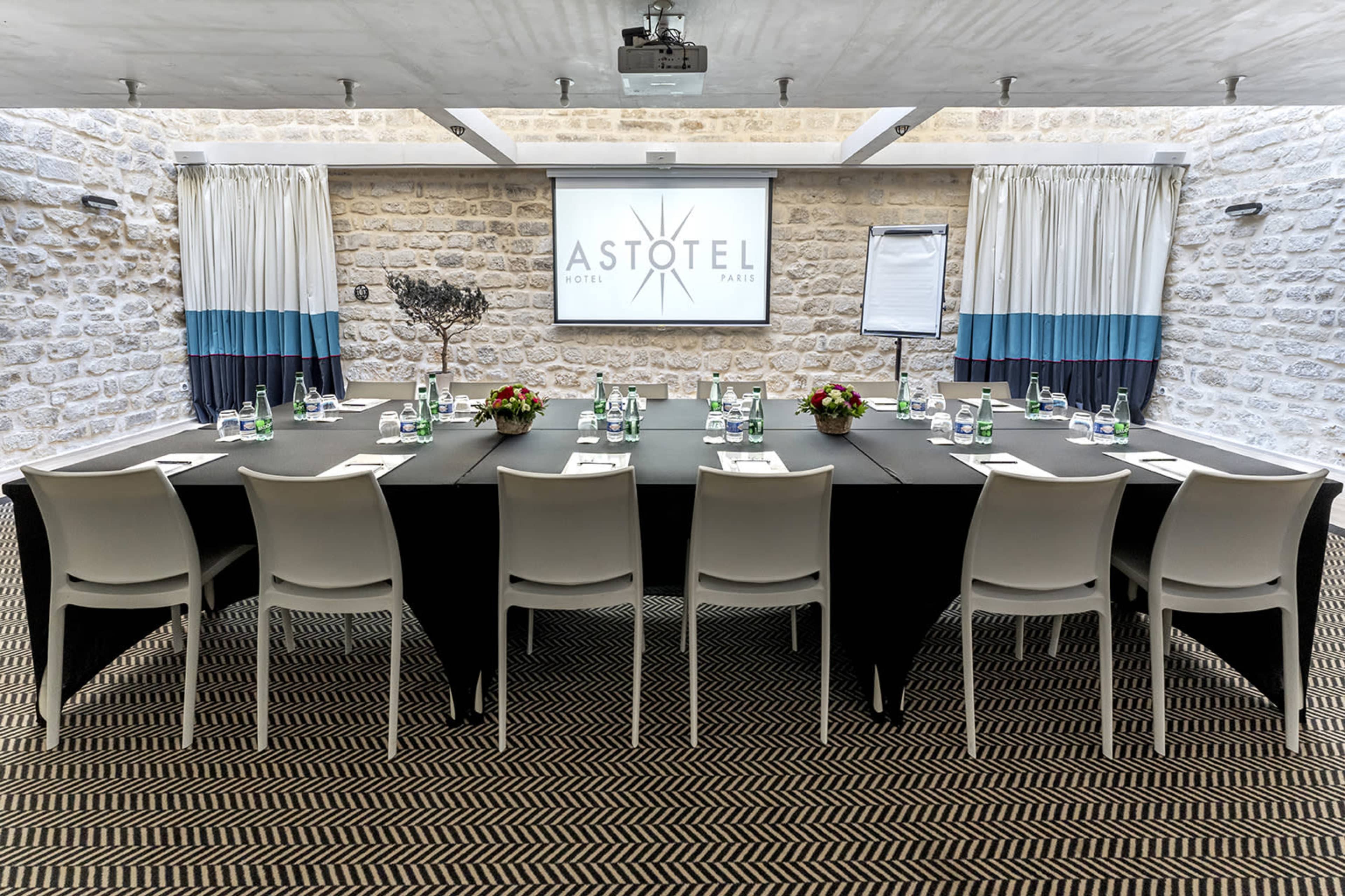 A conference room is set up with a long table, chairs, water bottles, and floral centerpieces, featuring a projection screen at the front.