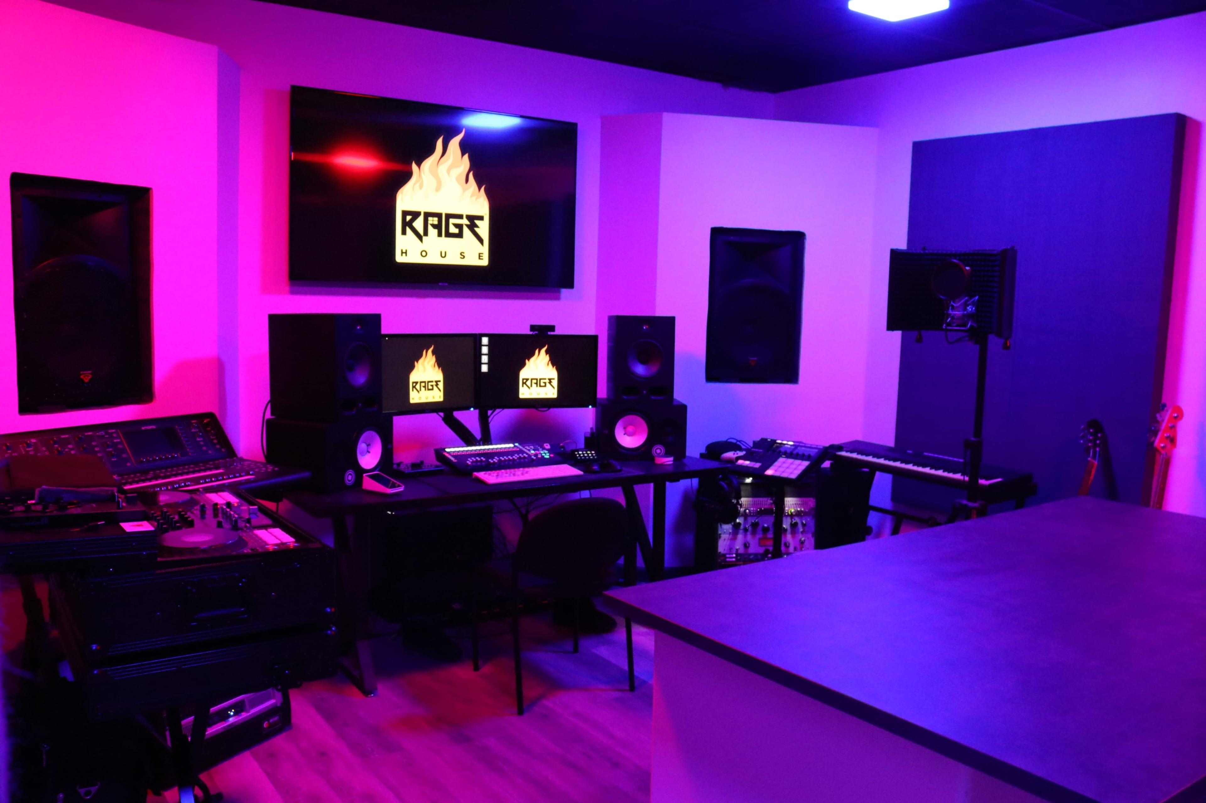 A music studio equipped with audio equipment, two computer monitors, speakers, and soundproofing panels, illuminated by purple and pink lighting.