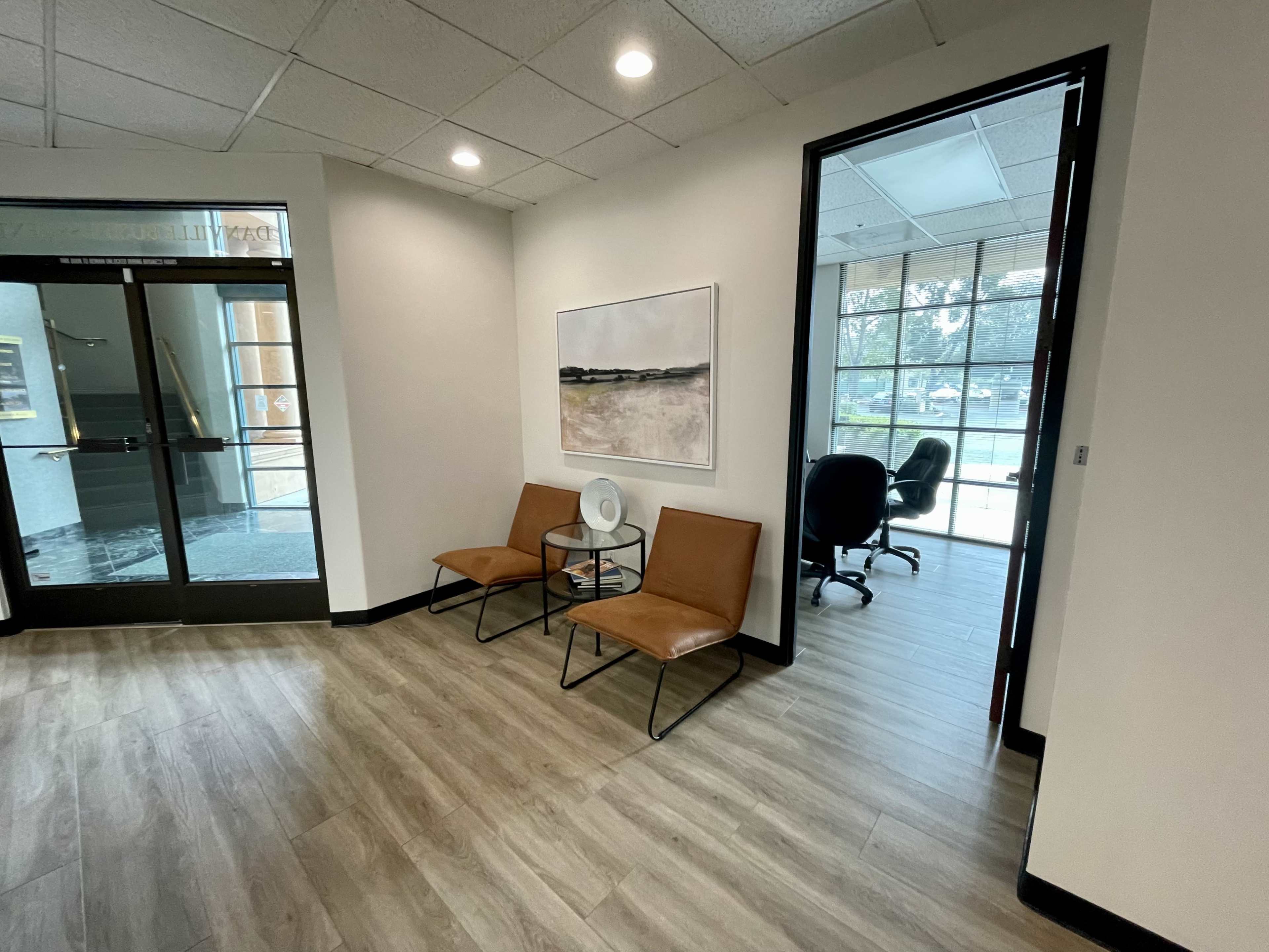 A modern office entryway with two brown chairs, a small fan, and a door leading to a workspace filled with large windows.