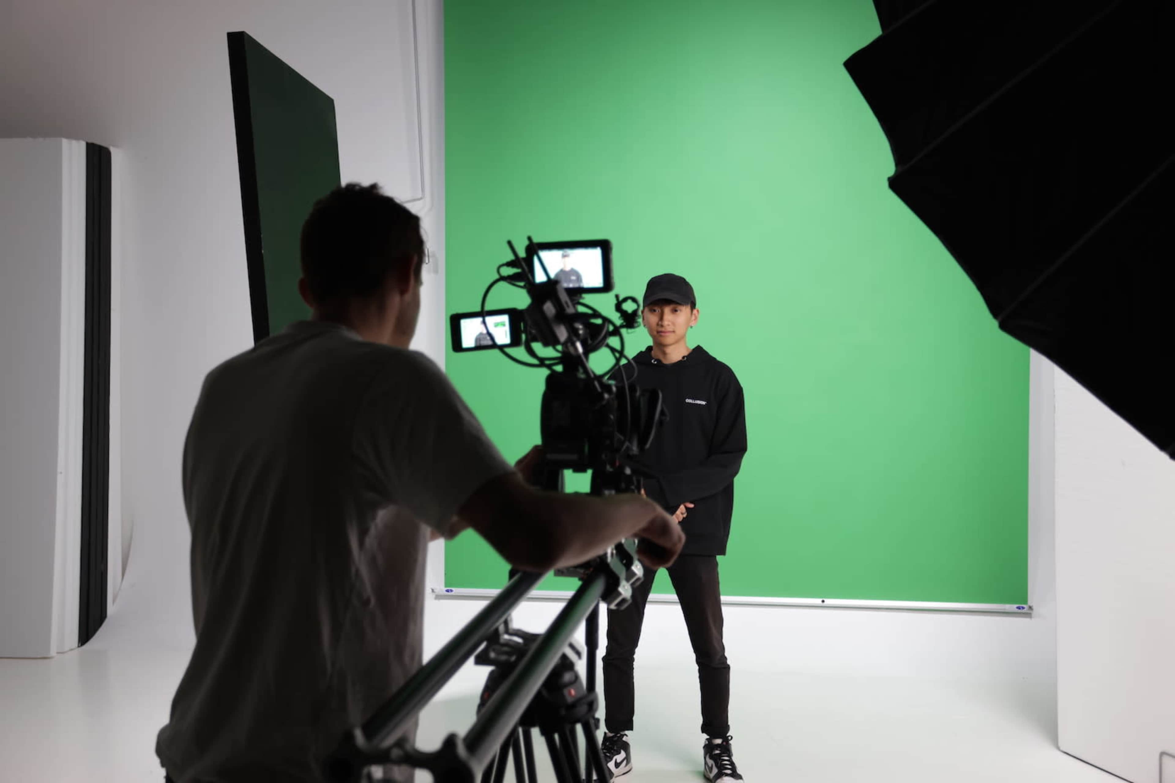 A person stands in front of a green screen while being filmed by a camera operator.