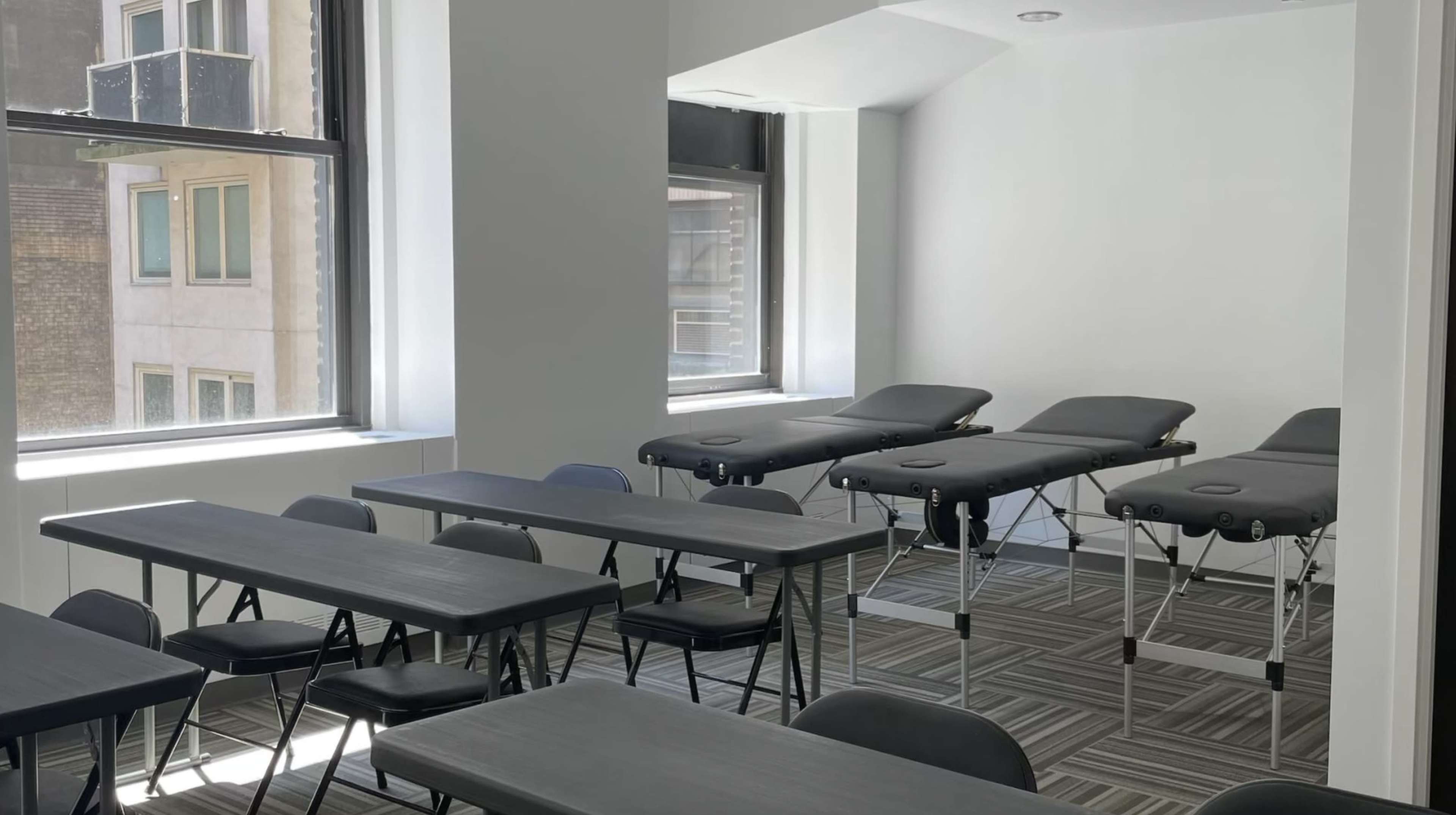 The image shows a room with two portable massage tables placed against a wall, surrounded by several chairs and tables arranged for a meeting or training session.