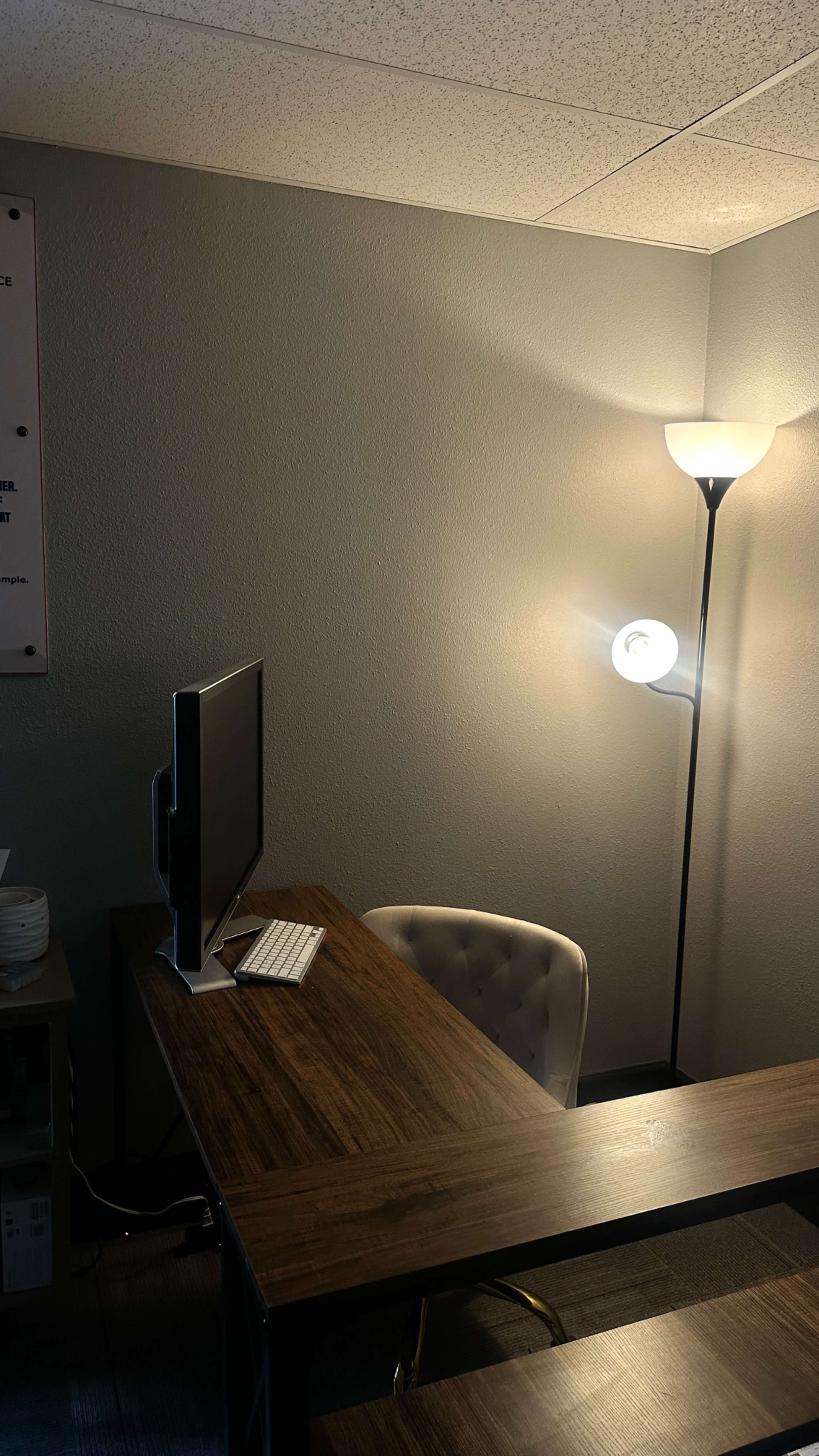 The image shows a minimalistic office space featuring a wooden desk with a computer and a chair, illuminated by a nearby lamp against a gray wall.