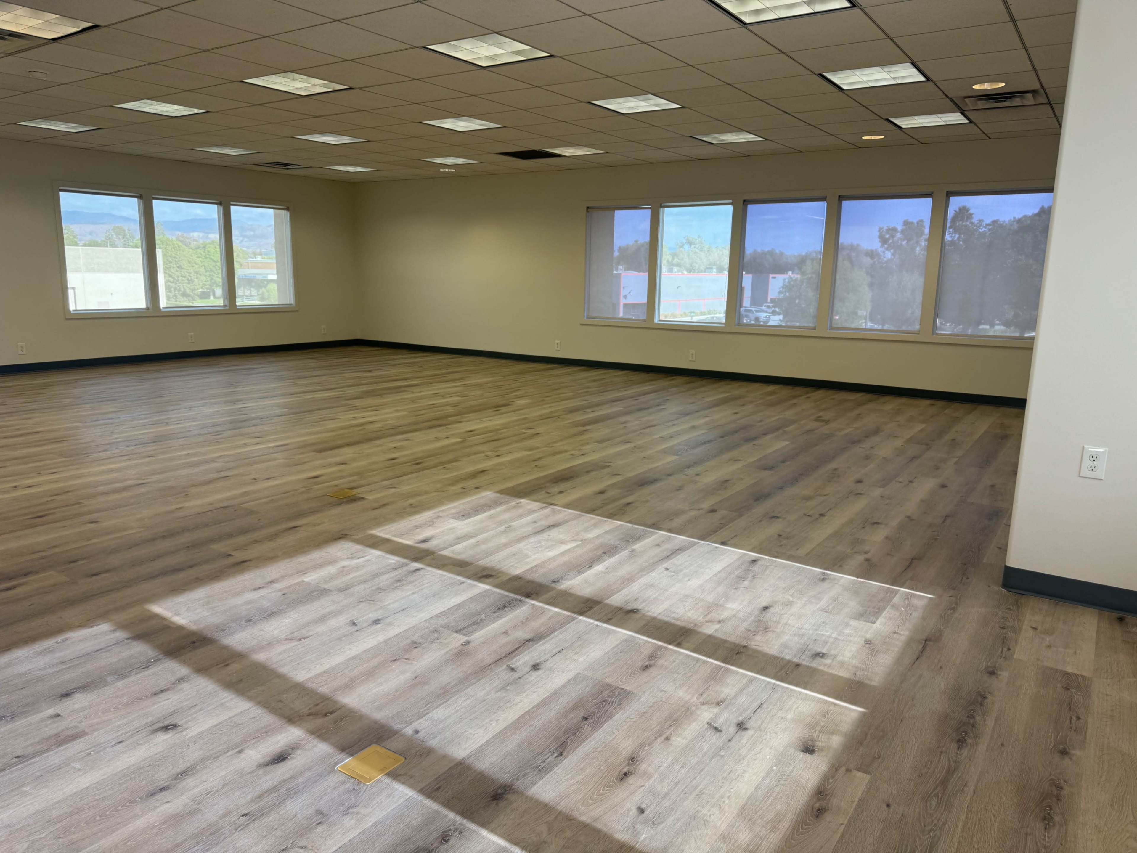 The image shows a spacious empty office room with wooden flooring and several large windows allowing natural light to enter.