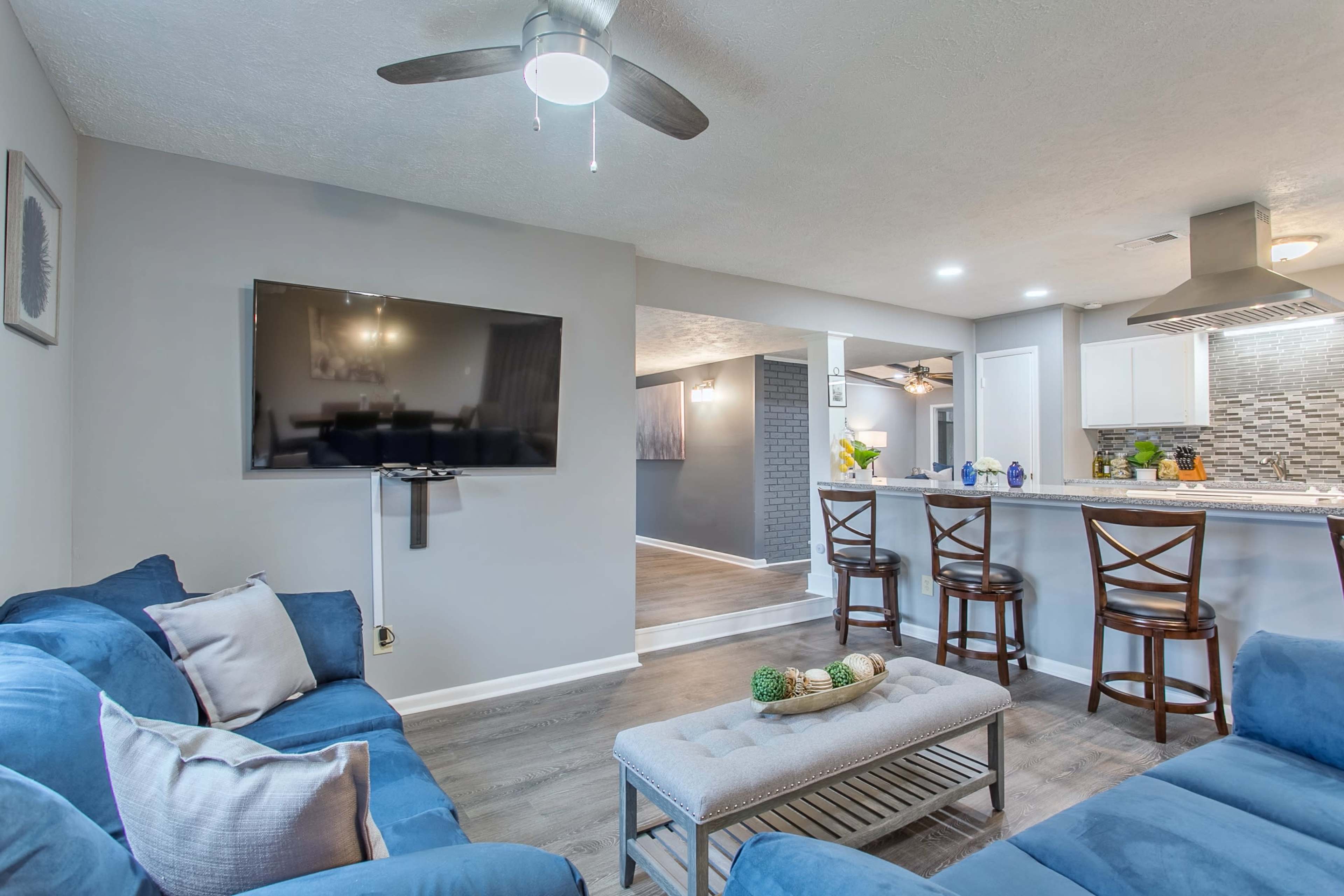 A living room with blue sofas, a mounted television, and an open layout leading to a kitchen with bar seating and modern decor.