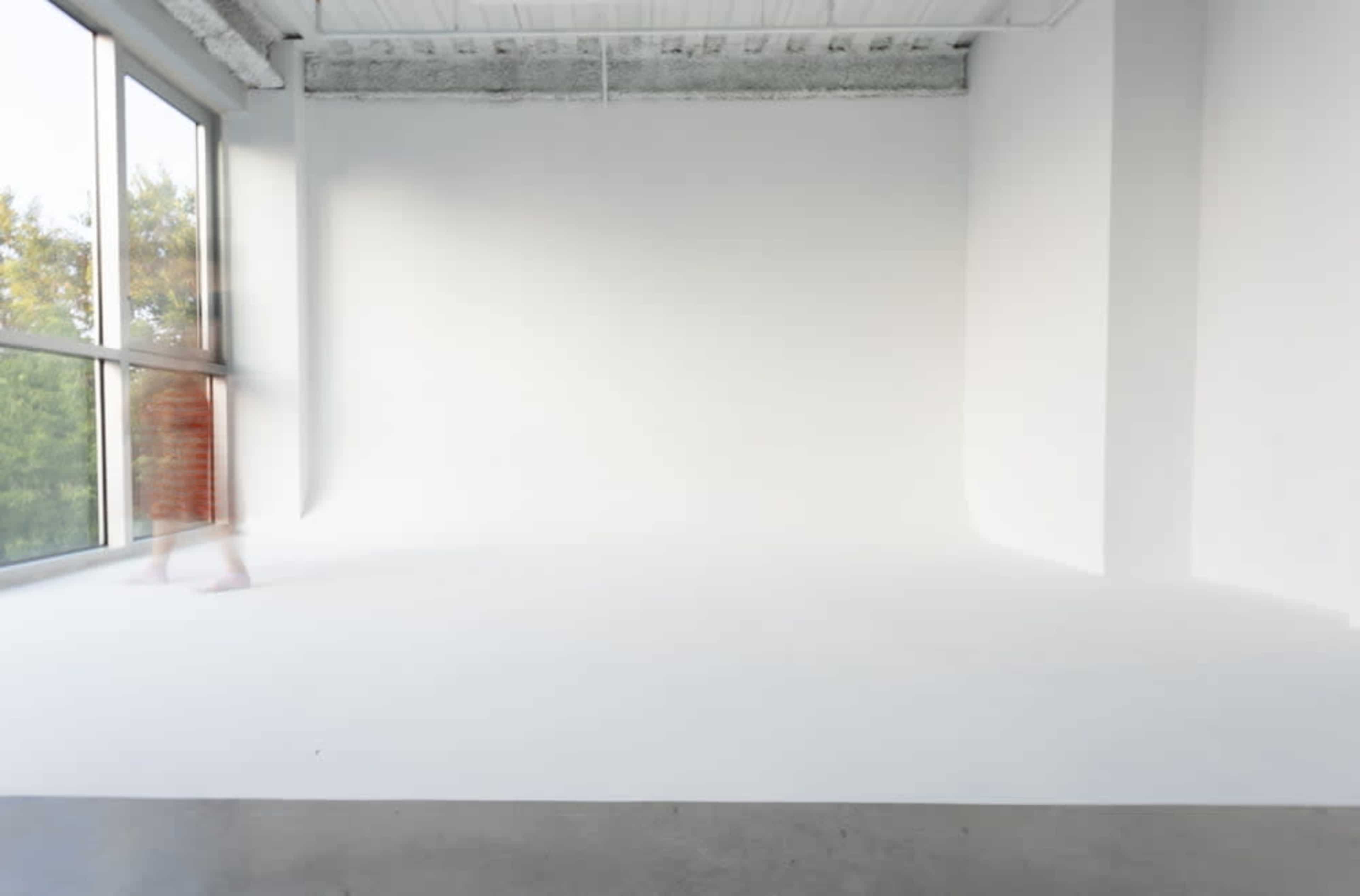 A person walks through an empty, well-lit room with white walls and a large window.