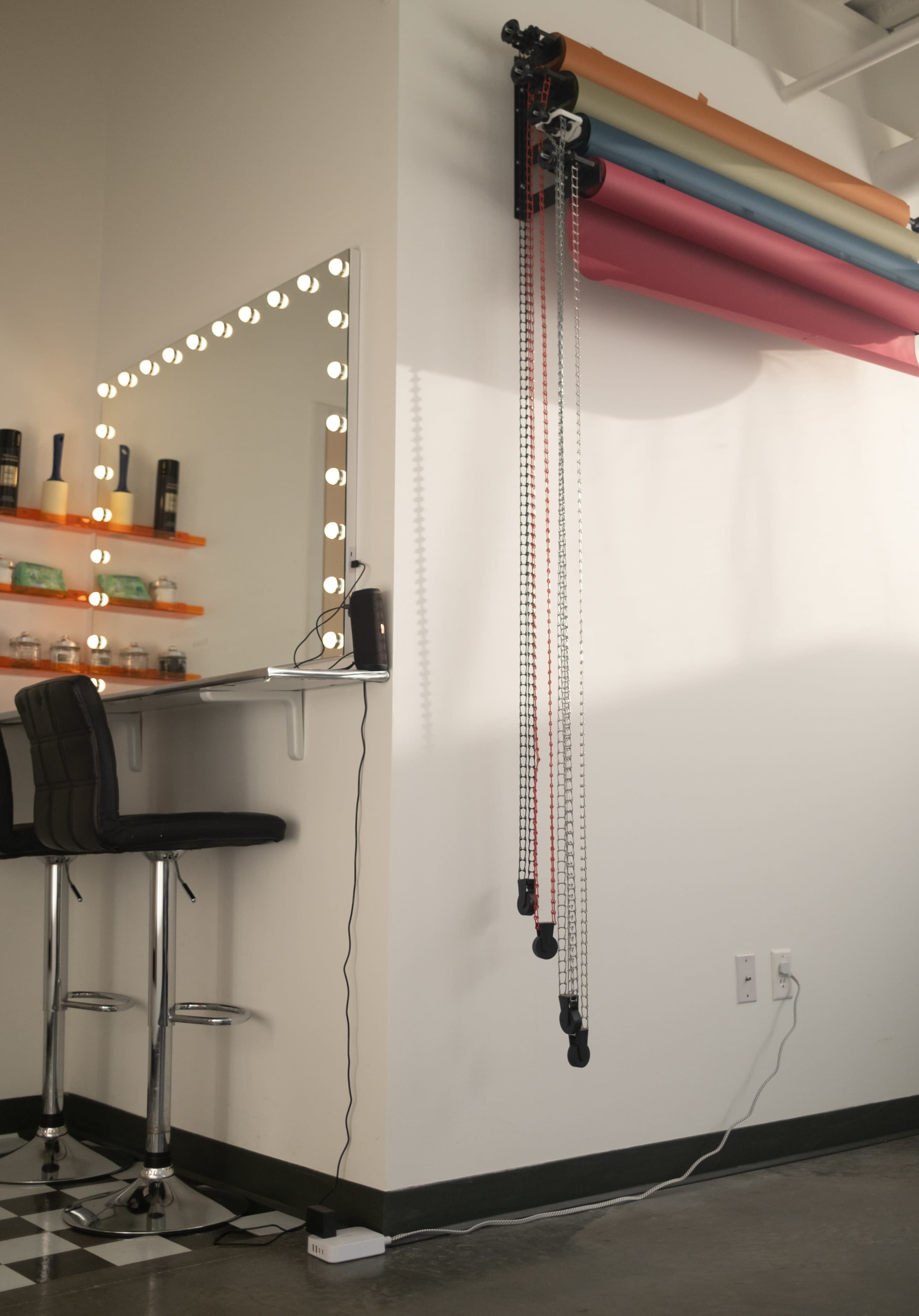 A makeup vanity with a lit mirror stands beside a wall that features a multicolored backdrop hung on a rolling system.