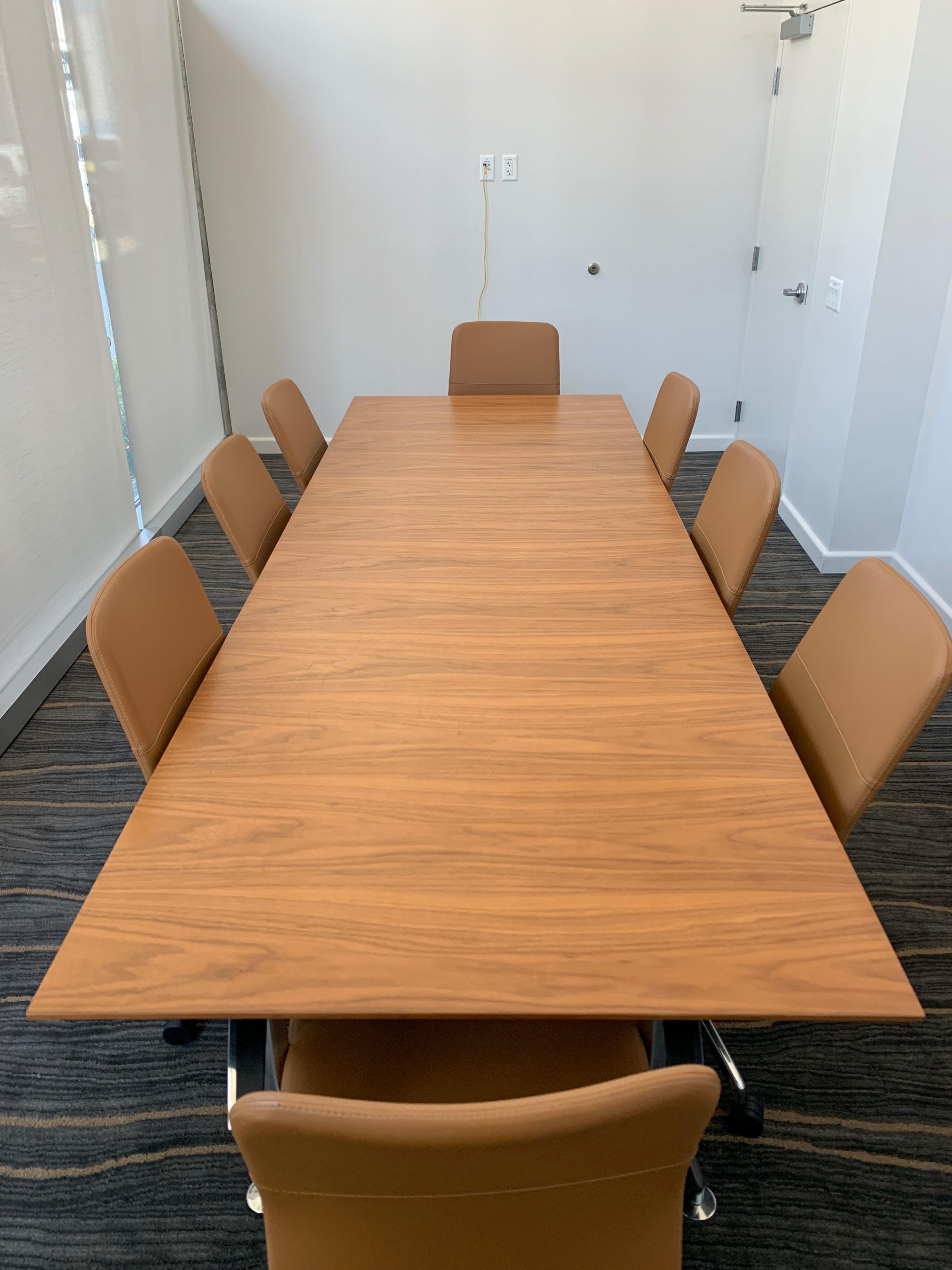 A rectangular wooden conference table is surrounded by eight brown chairs in a well-lit meeting room.