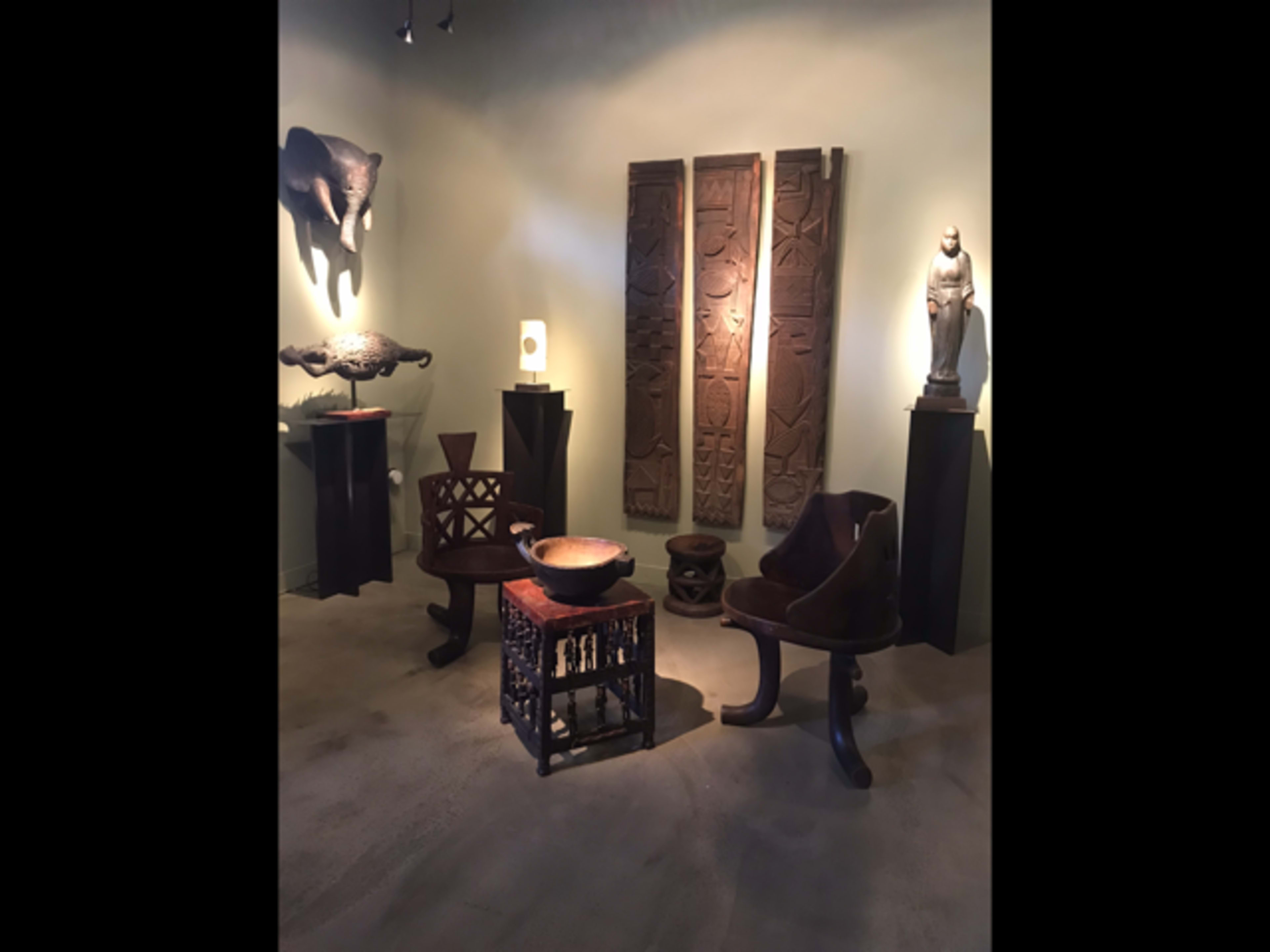 An art gallery displaying various wooden sculptures and artifacts, including an elephant figure, carved panels, and furniture pieces arranged in a softly lit space.