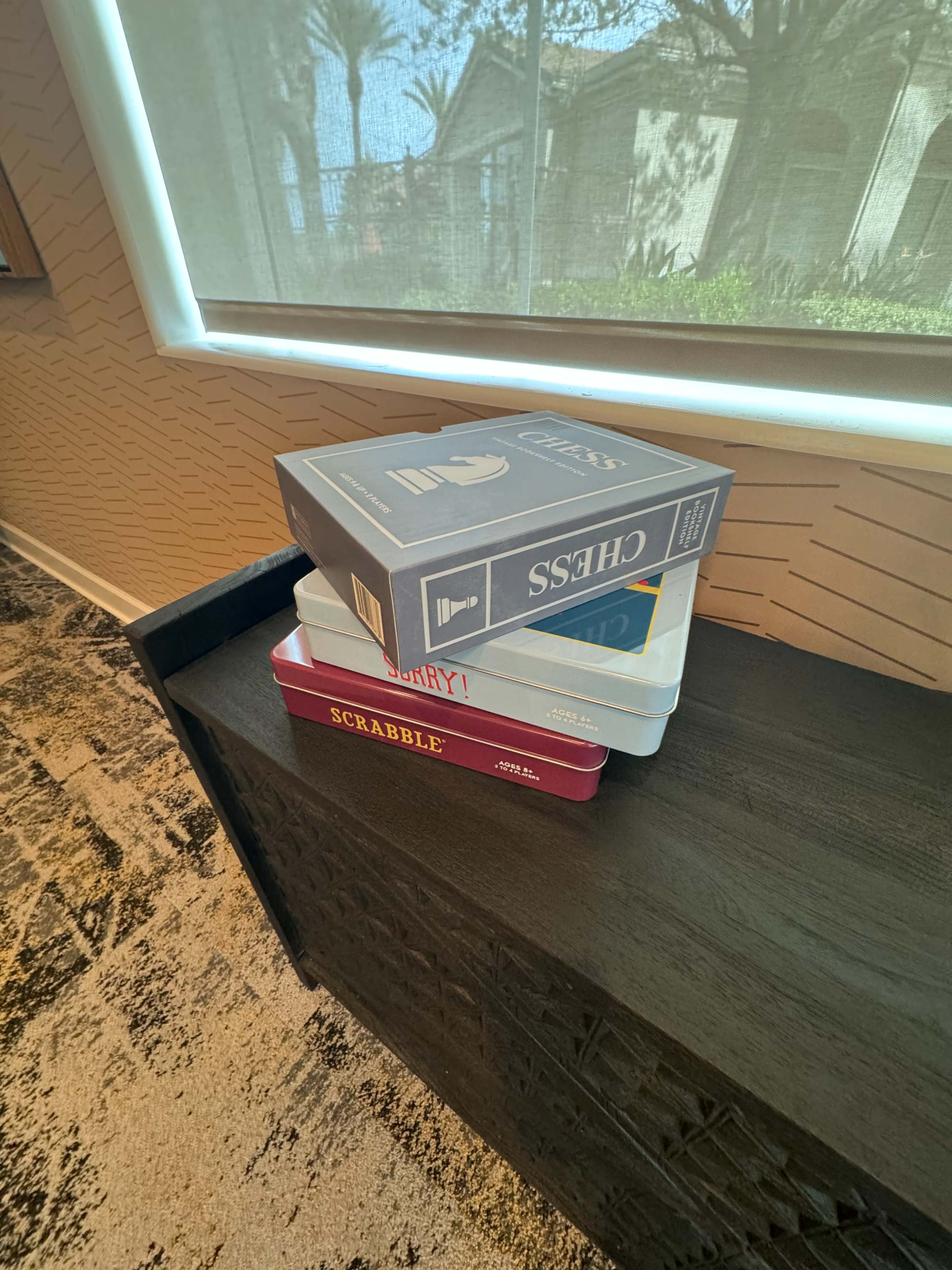 A stack of board game boxes, including Scrabble, Sorry, and Chess, rests on a dark wooden surface near a window.