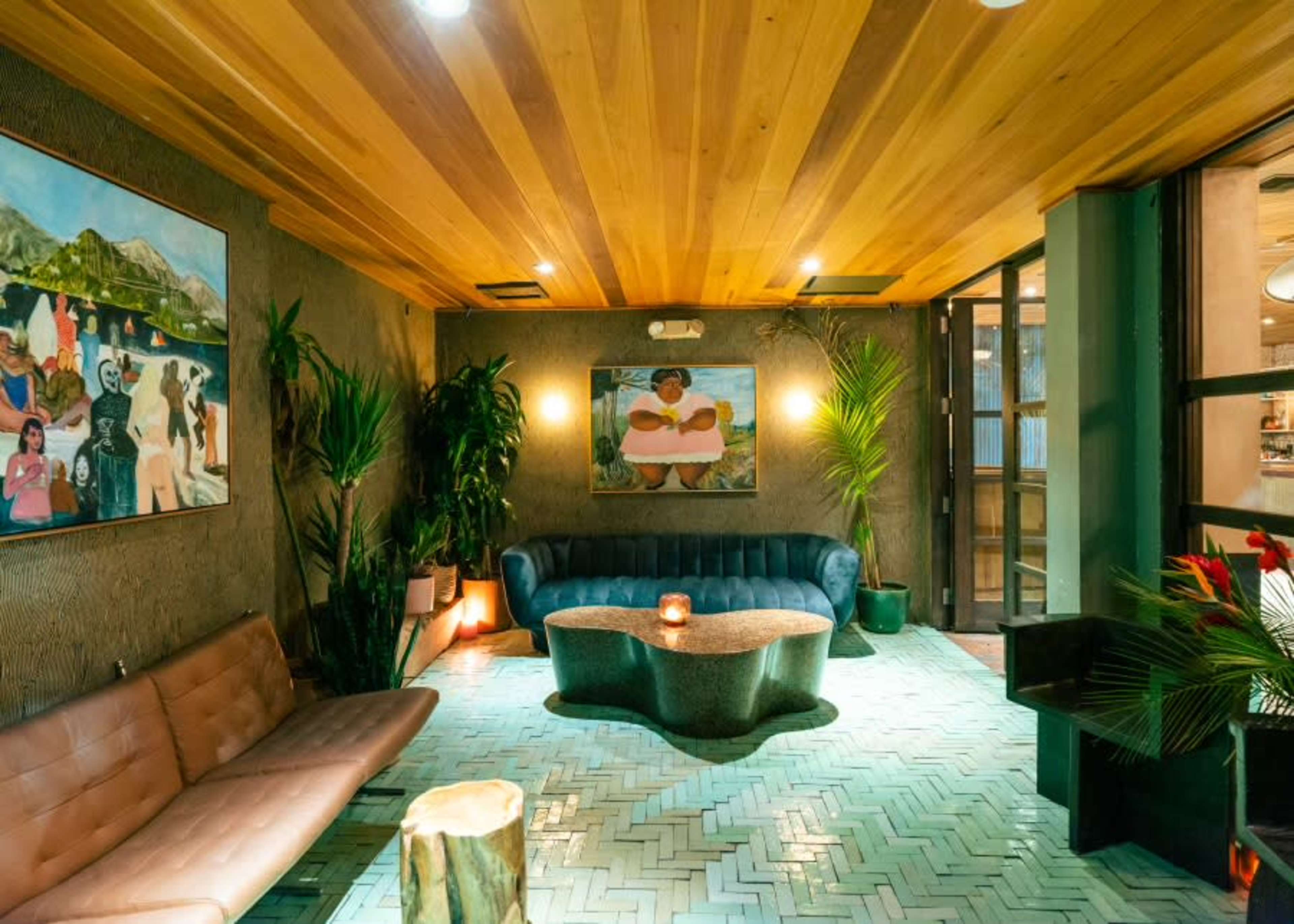The image shows a stylish lounge area featuring a blue couch, a wood coffee table, and tropical plants, with art on the walls.