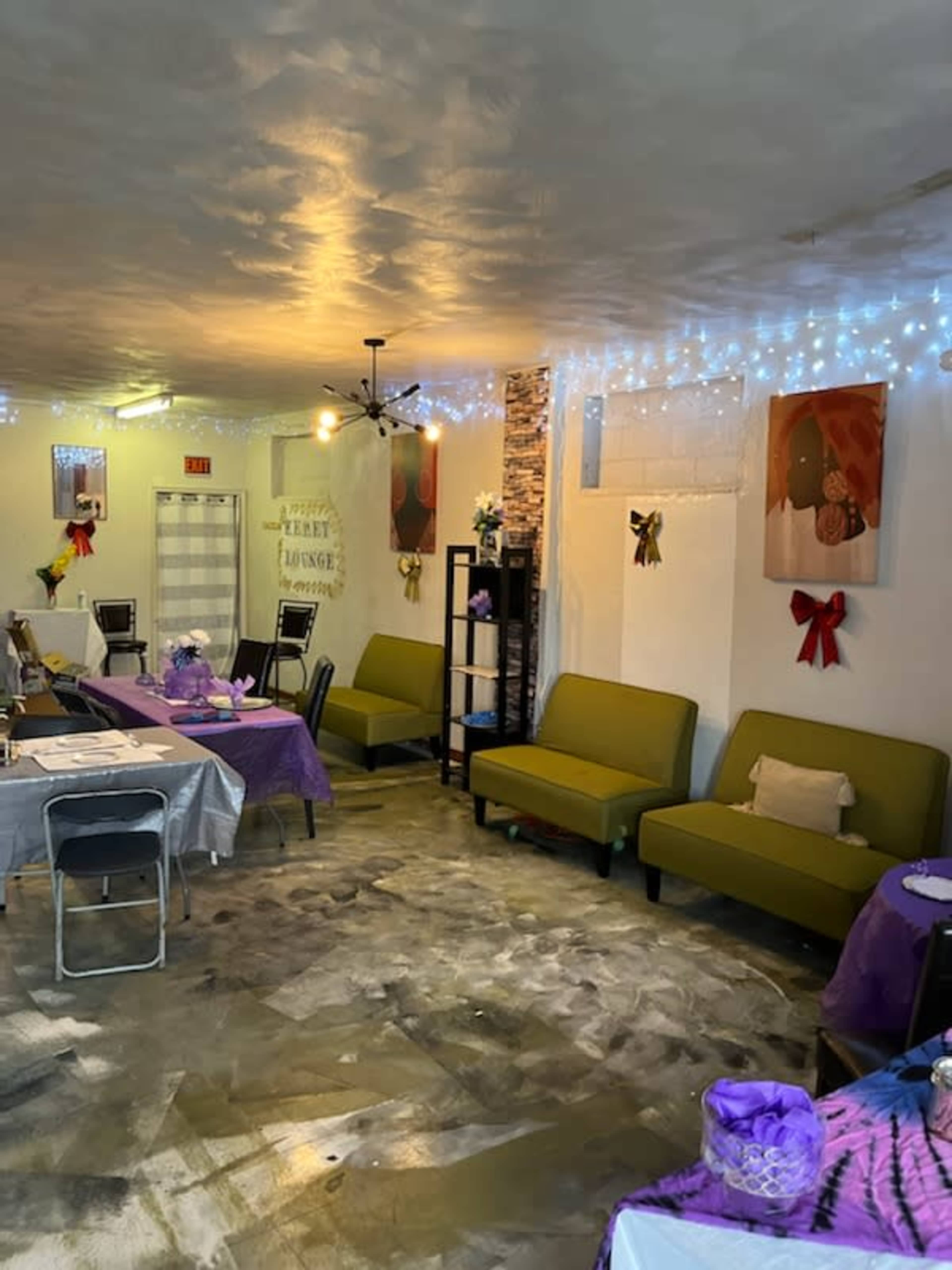 The image shows an interior of a small cafe or event space, with green couches, tables covered in purple and gray tablecloths, and decorative lights on the walls.