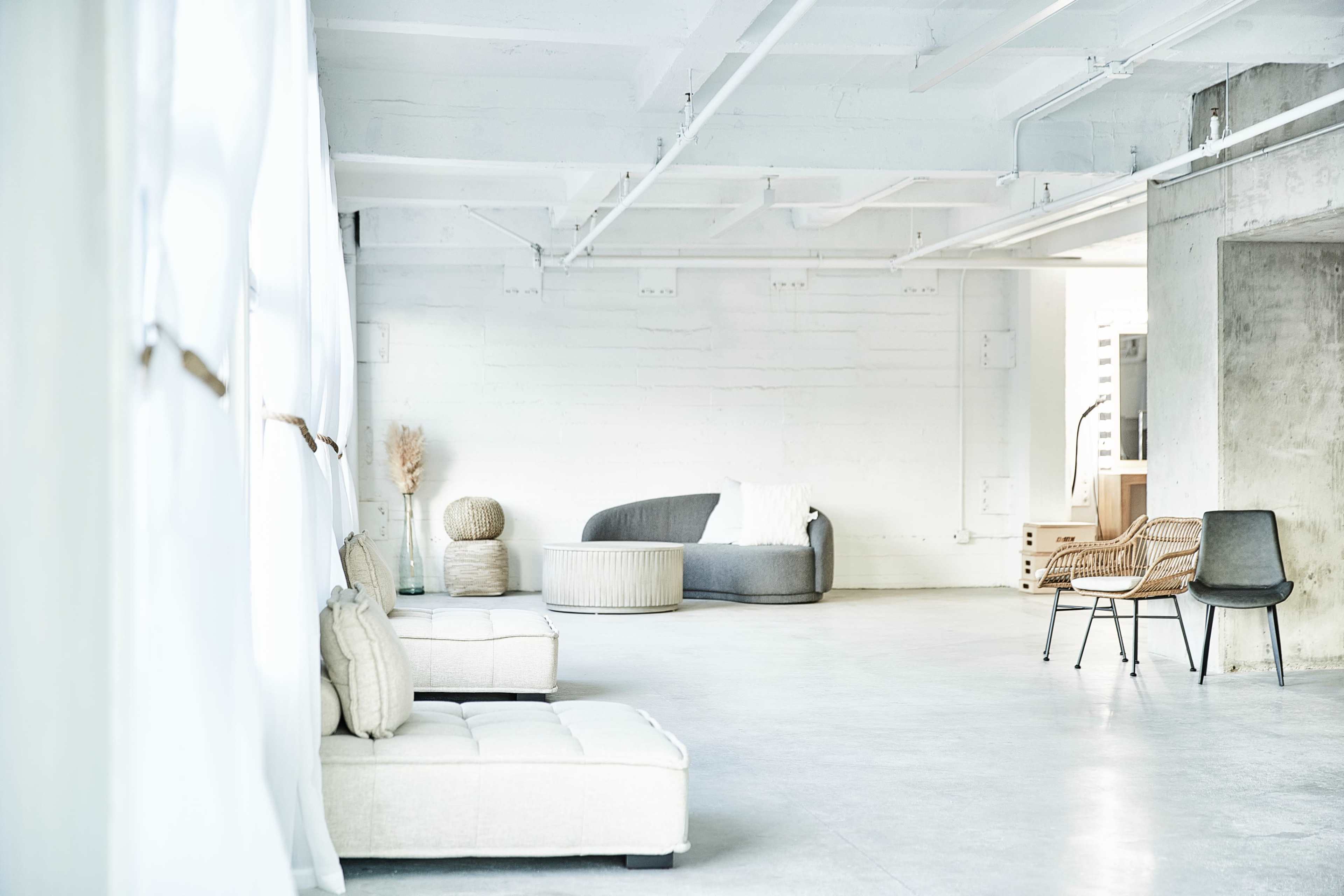 A spacious, minimalist interior with various pieces of furniture, including a gray sofa, a round ottoman, and several chairs, set against a light-colored, industrial wall.