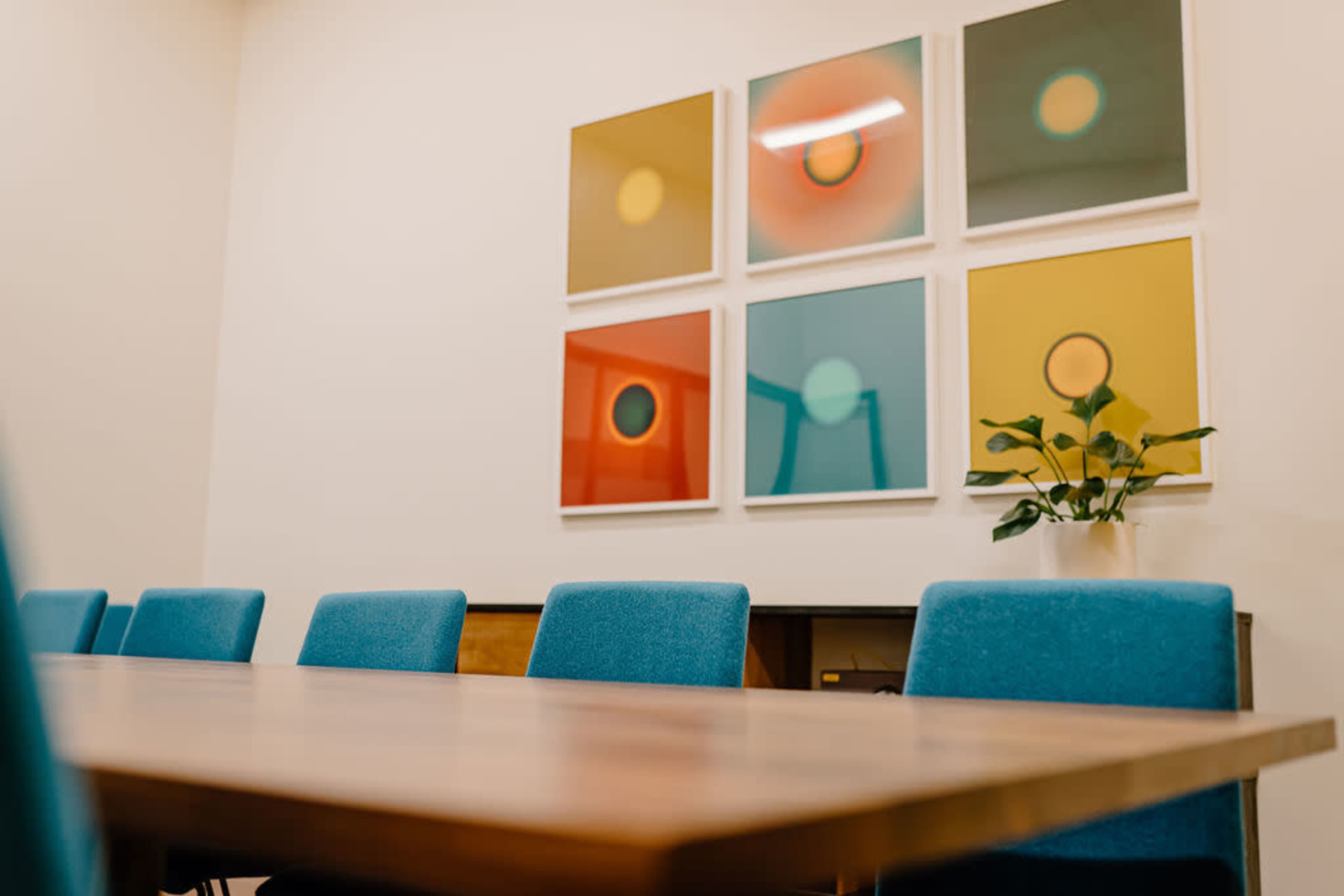 A conference room features a wooden table surrounded by blue chairs and colorful framed artwork on the wall.