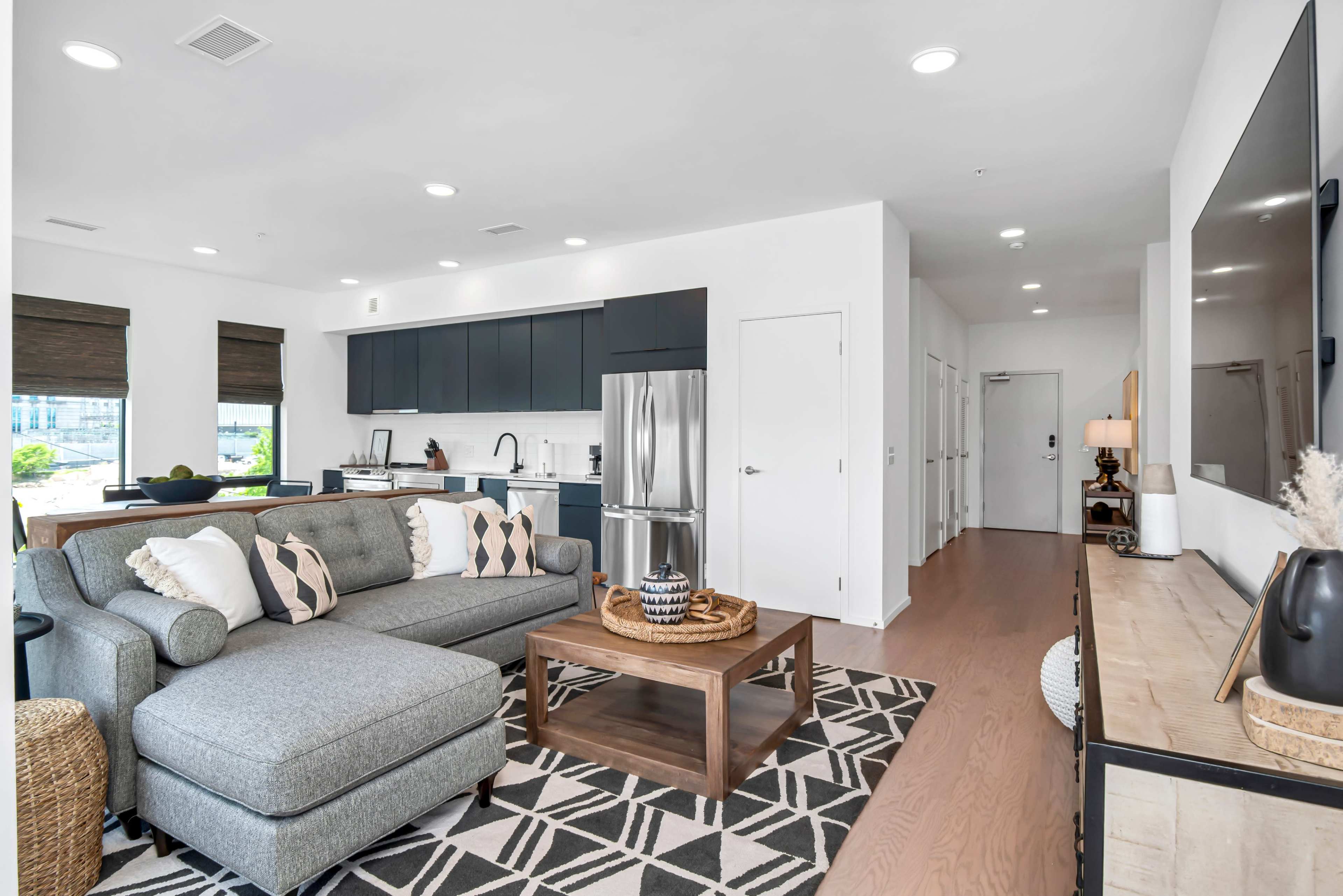 A modern living room and kitchen area featuring a gray sofa, a coffee table, and a kitchen with dark cabinets and stainless steel appliances.
