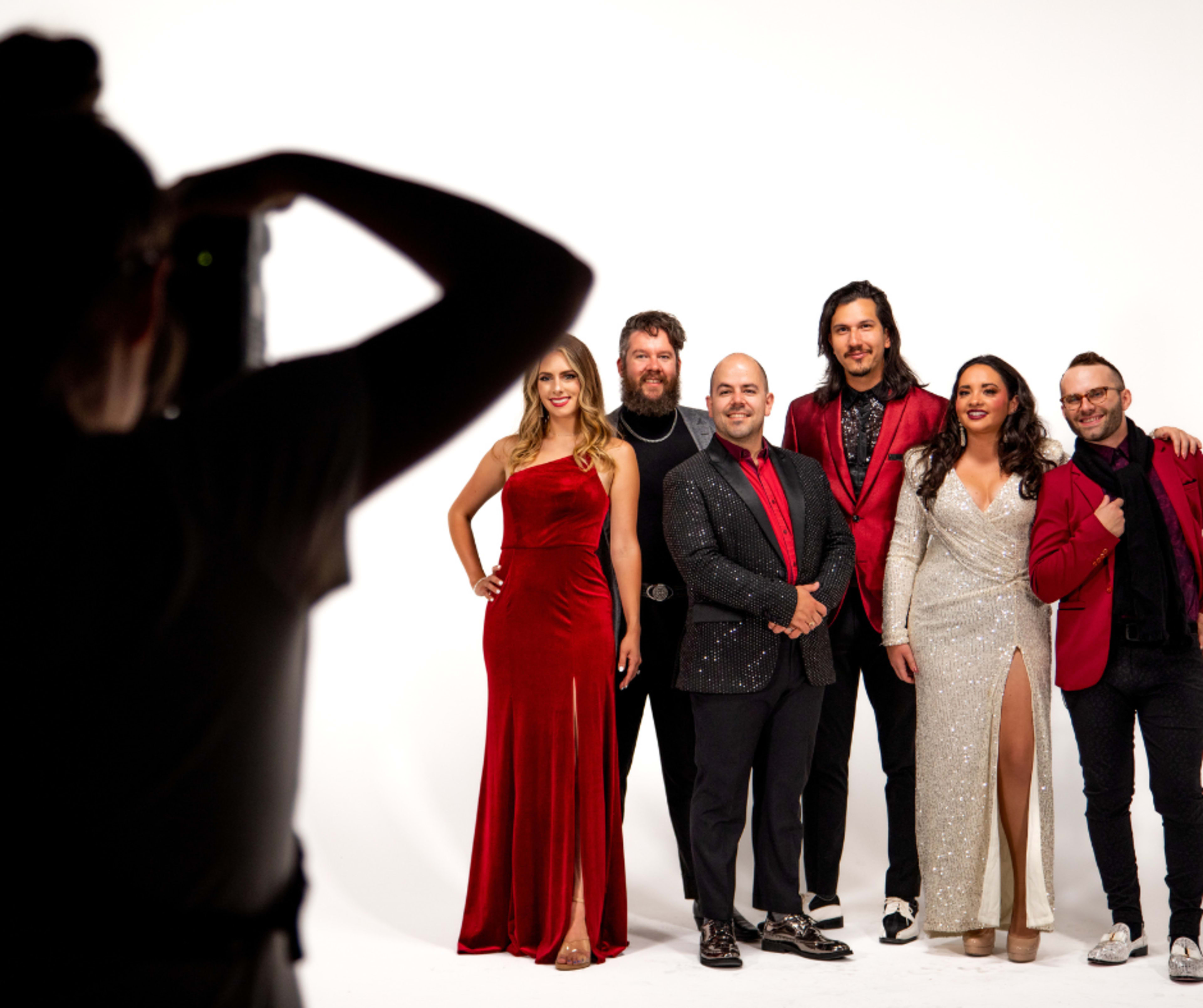 A group of six people poses for a photograph against a white backdrop, with one person holding a camera.