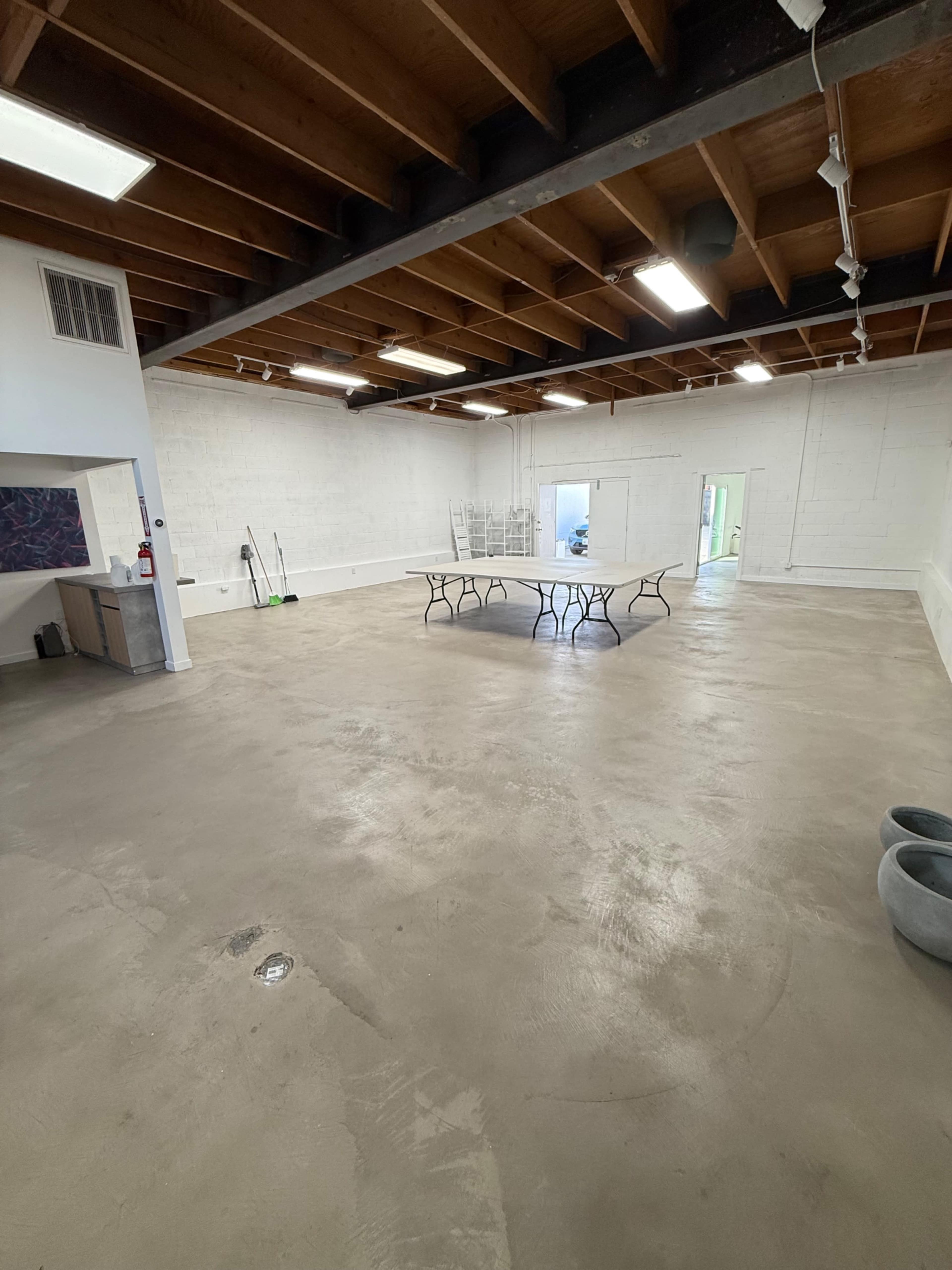 The image shows an empty, spacious room with a concrete floor, exposed wooden beams on the ceiling, and a central table surrounded by chairs.