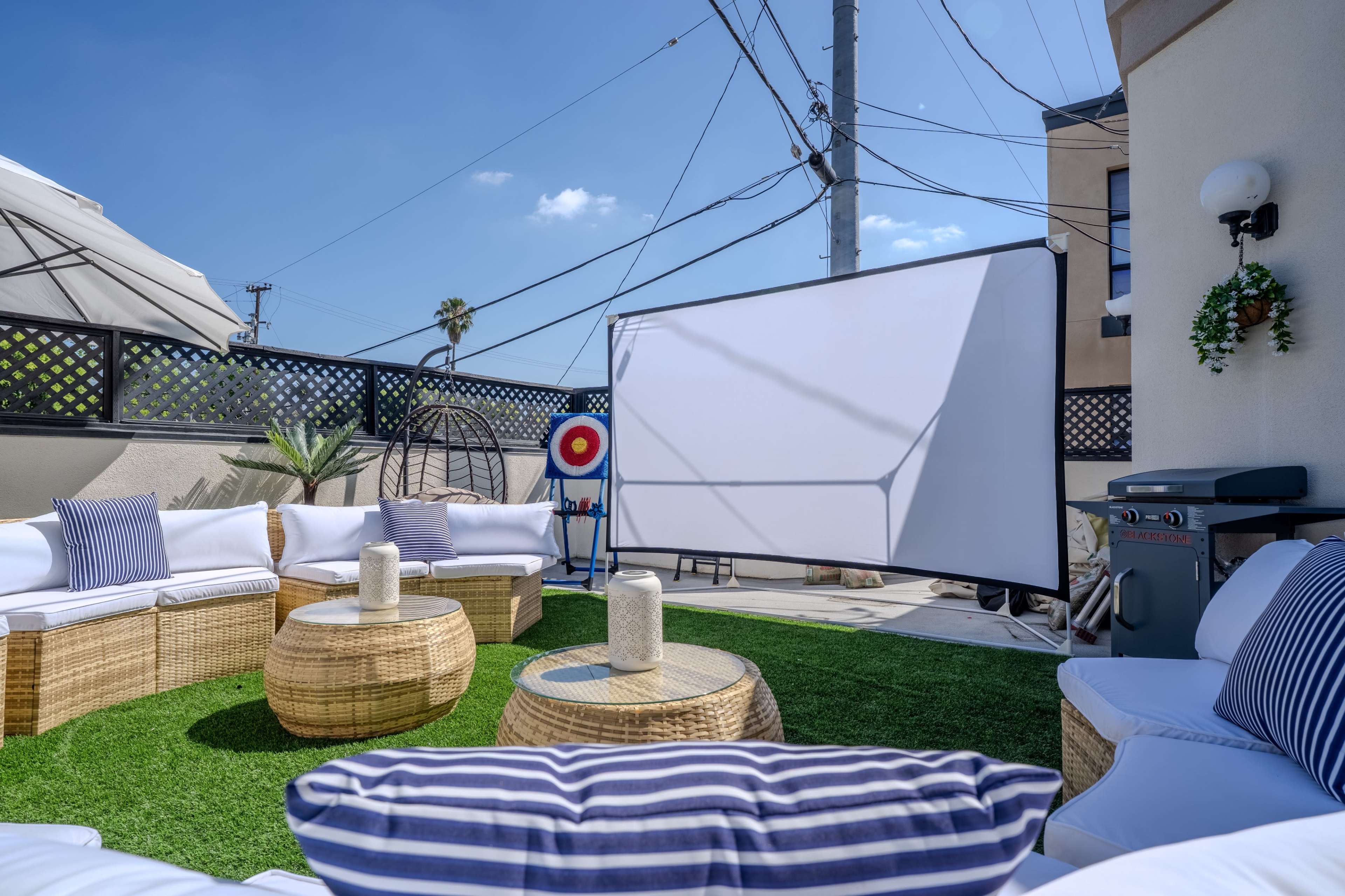 The image shows a patio area with outdoor seating, a large projection screen, a barbecue grill, and power lines in the background under a clear blue sky.