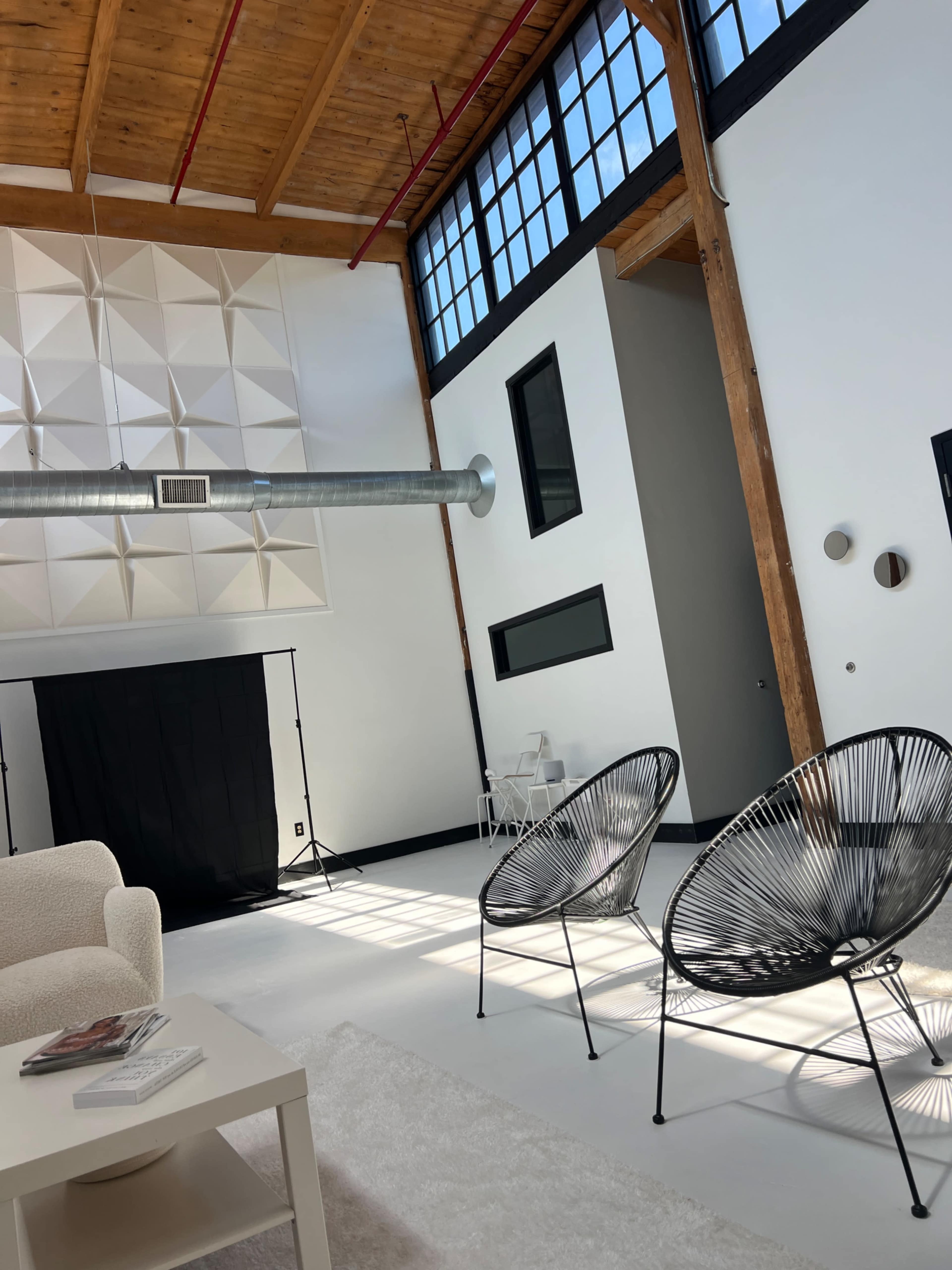 A spacious interior featuring a large window, a black backdrop, two wire chairs, and a light-colored sofa on a rug.