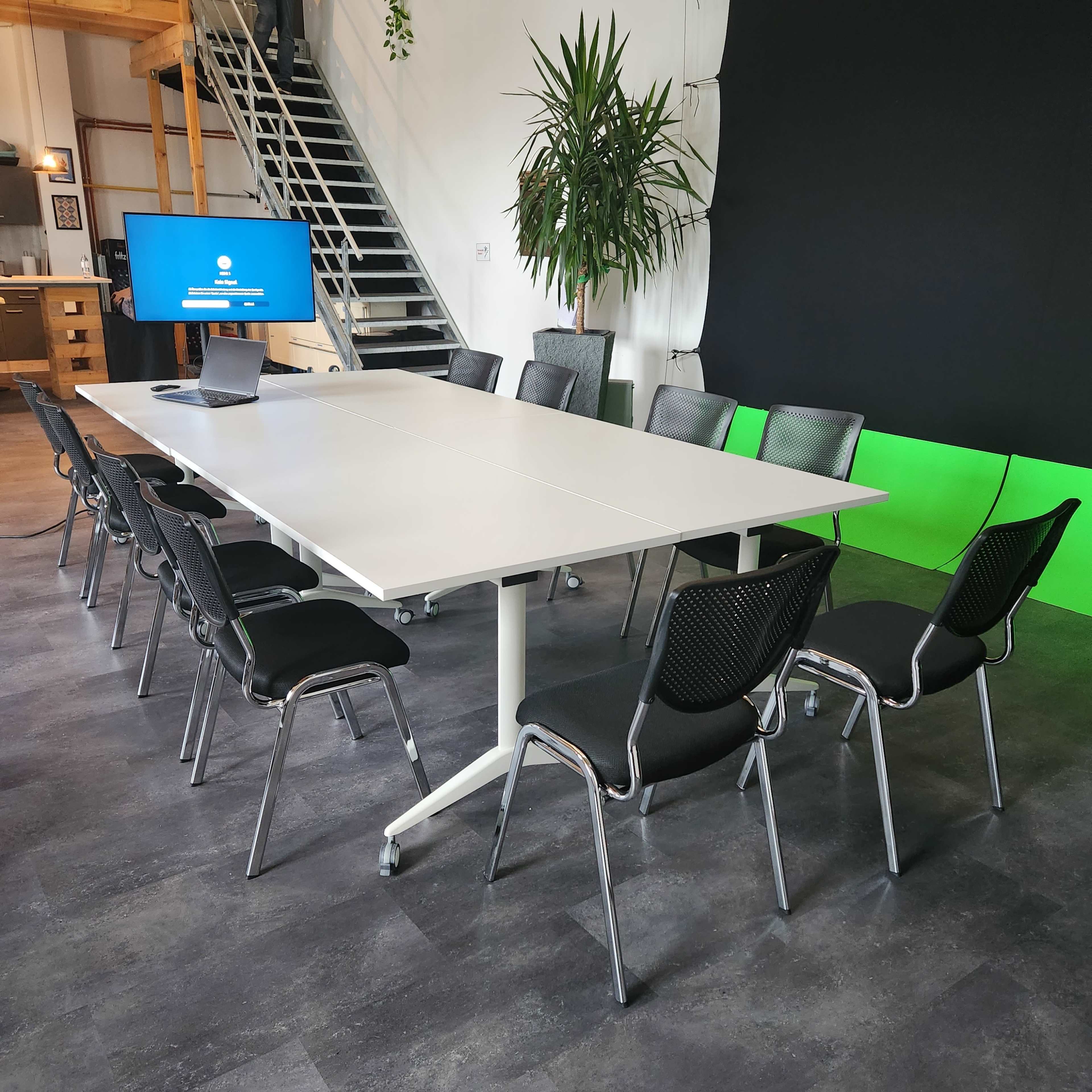 A modern conference room features a large white table surrounded by black chairs, with a television screen displaying information in the background.