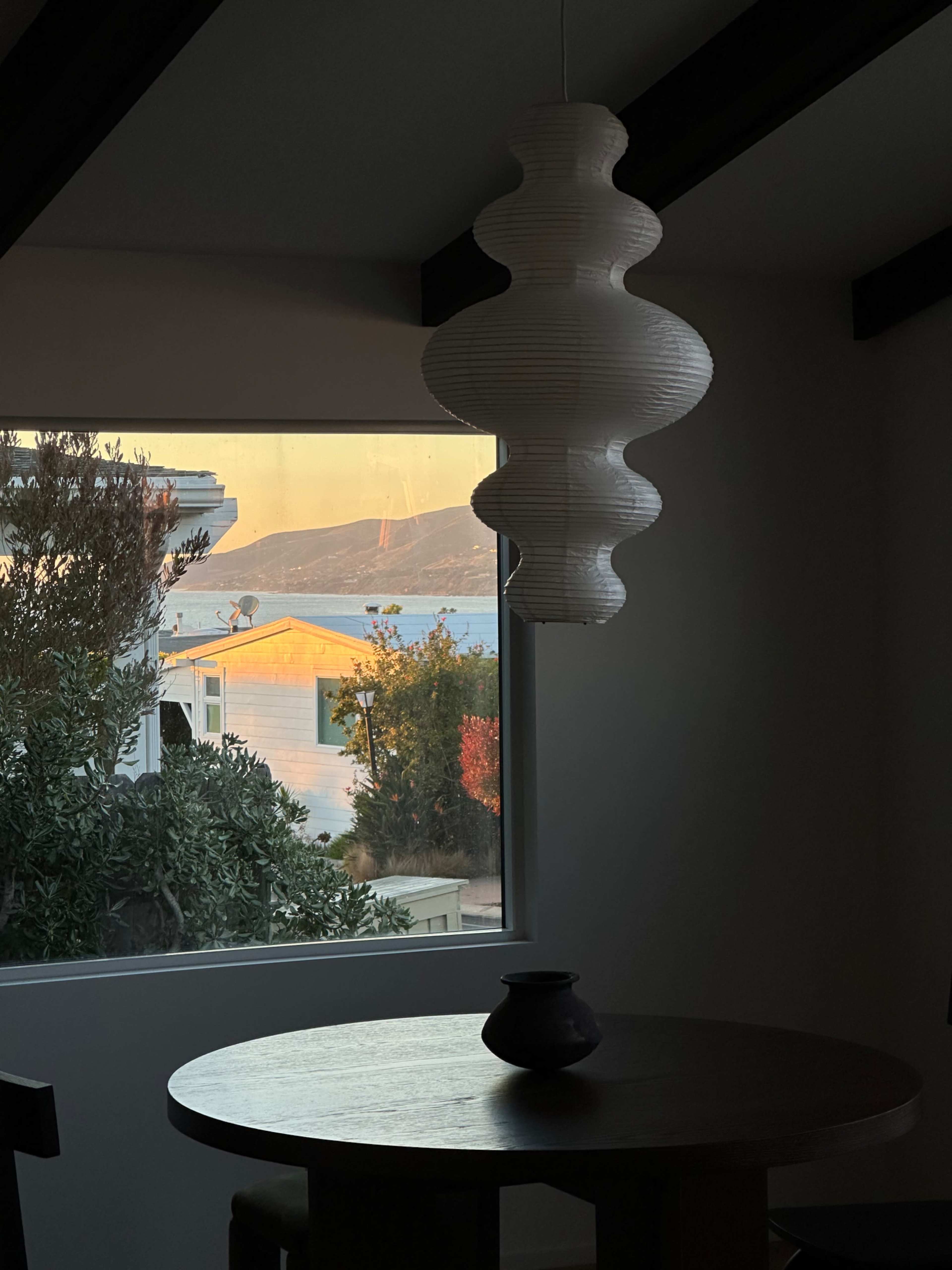 The image shows a dining table with a dark vase, illuminated by light from a window overlooking a landscape with a body of water and hills in the background.