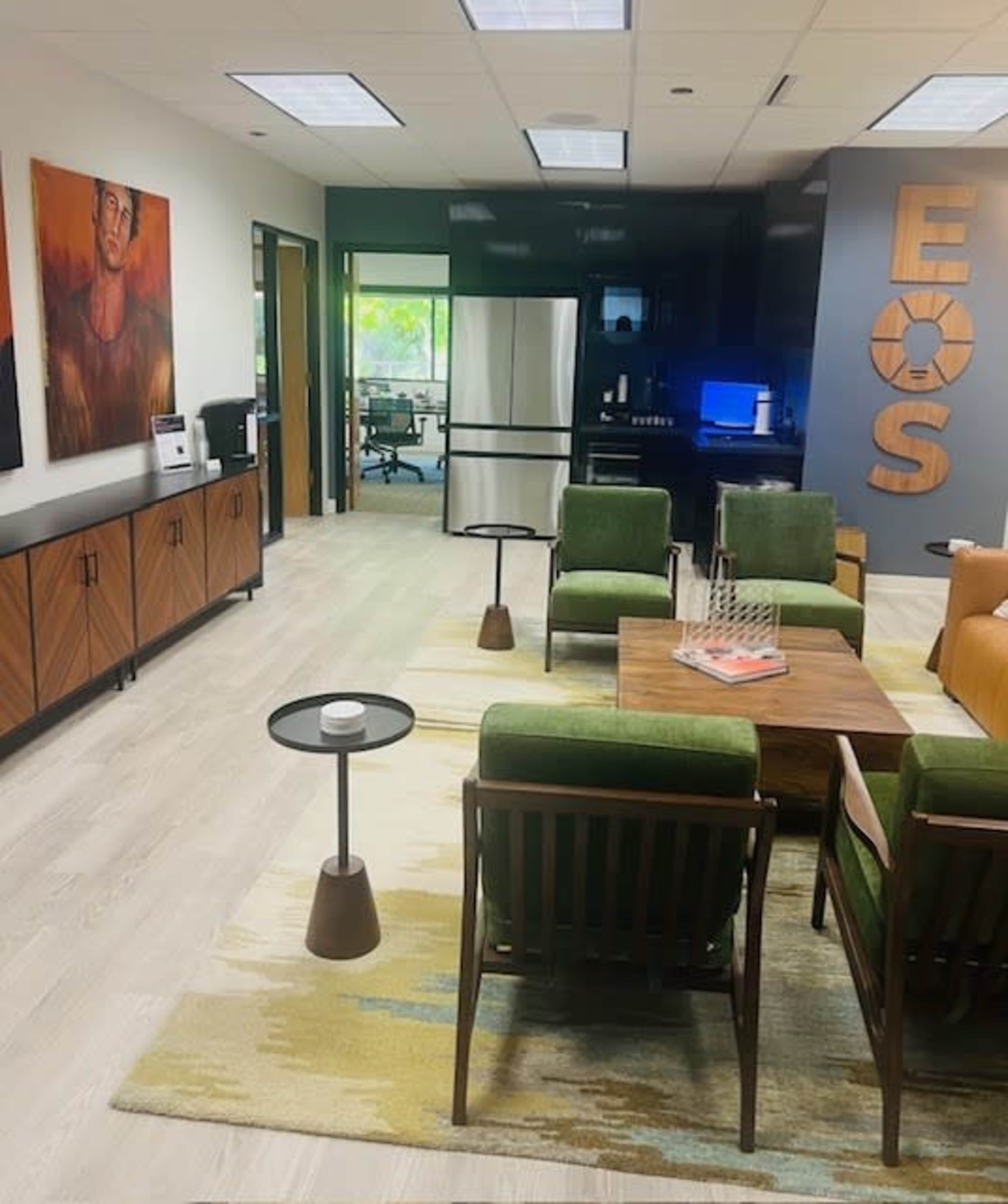 A modern office lounge area with green chairs, a wooden coffee table, and a decorative rug, alongside artwork on the walls and a kitchenette in the background.