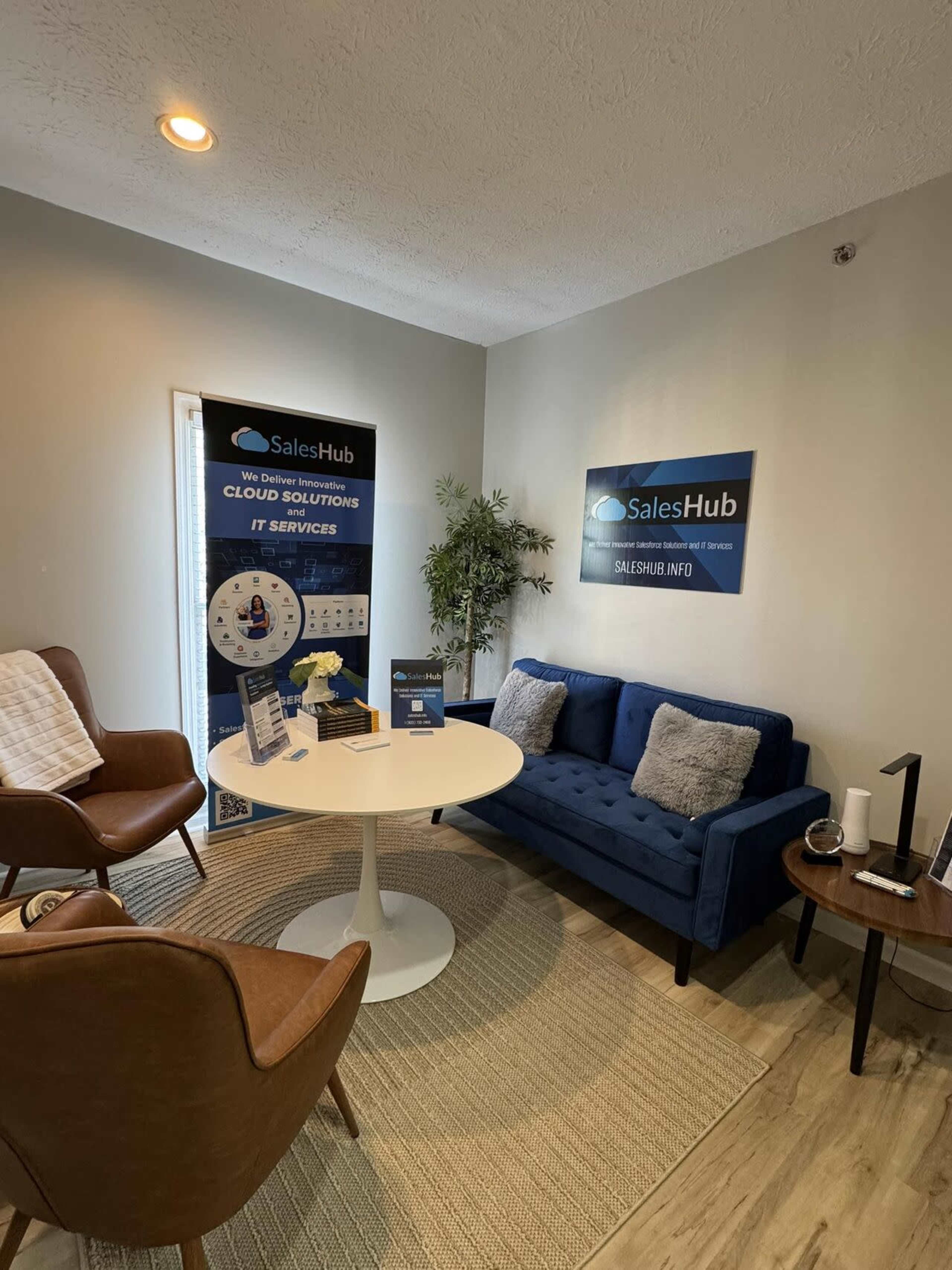 The image shows a small office waiting area with a round white table, blue sofa, brown chairs, promotional materials on the walls, and a decorative plant.