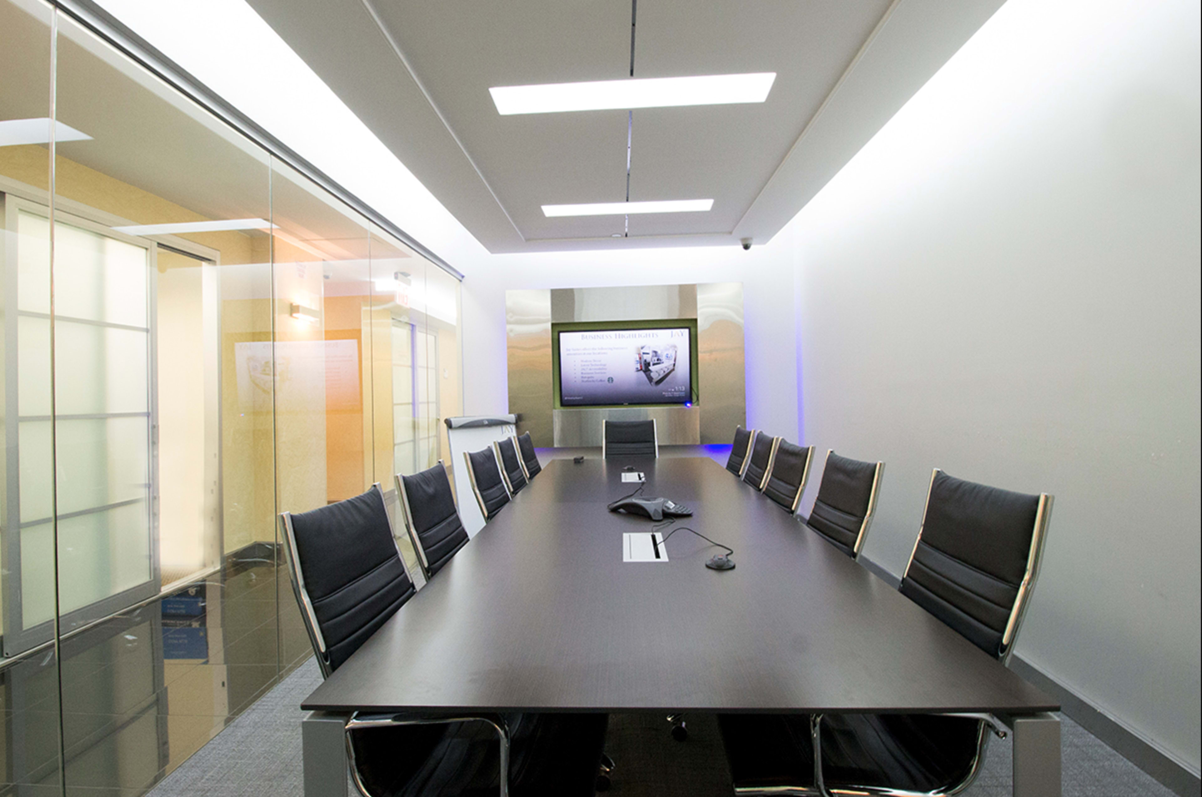 A modern conference room features a long table surrounded by black chairs, with a wall-mounted screen displaying information.
