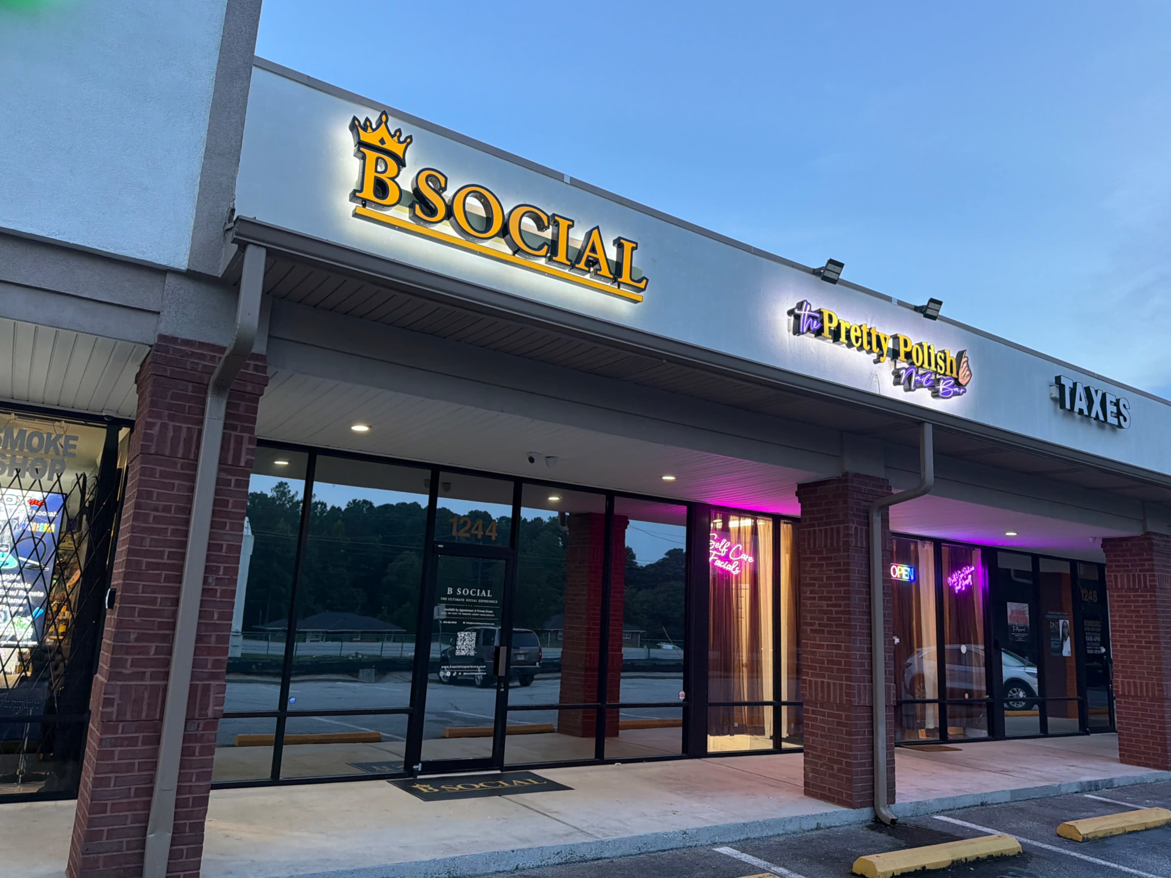 The image shows a shopping plaza with several storefronts, including "B SOCIAL," "Pretty Polish," and "TAXES," illuminated with signage and lights.