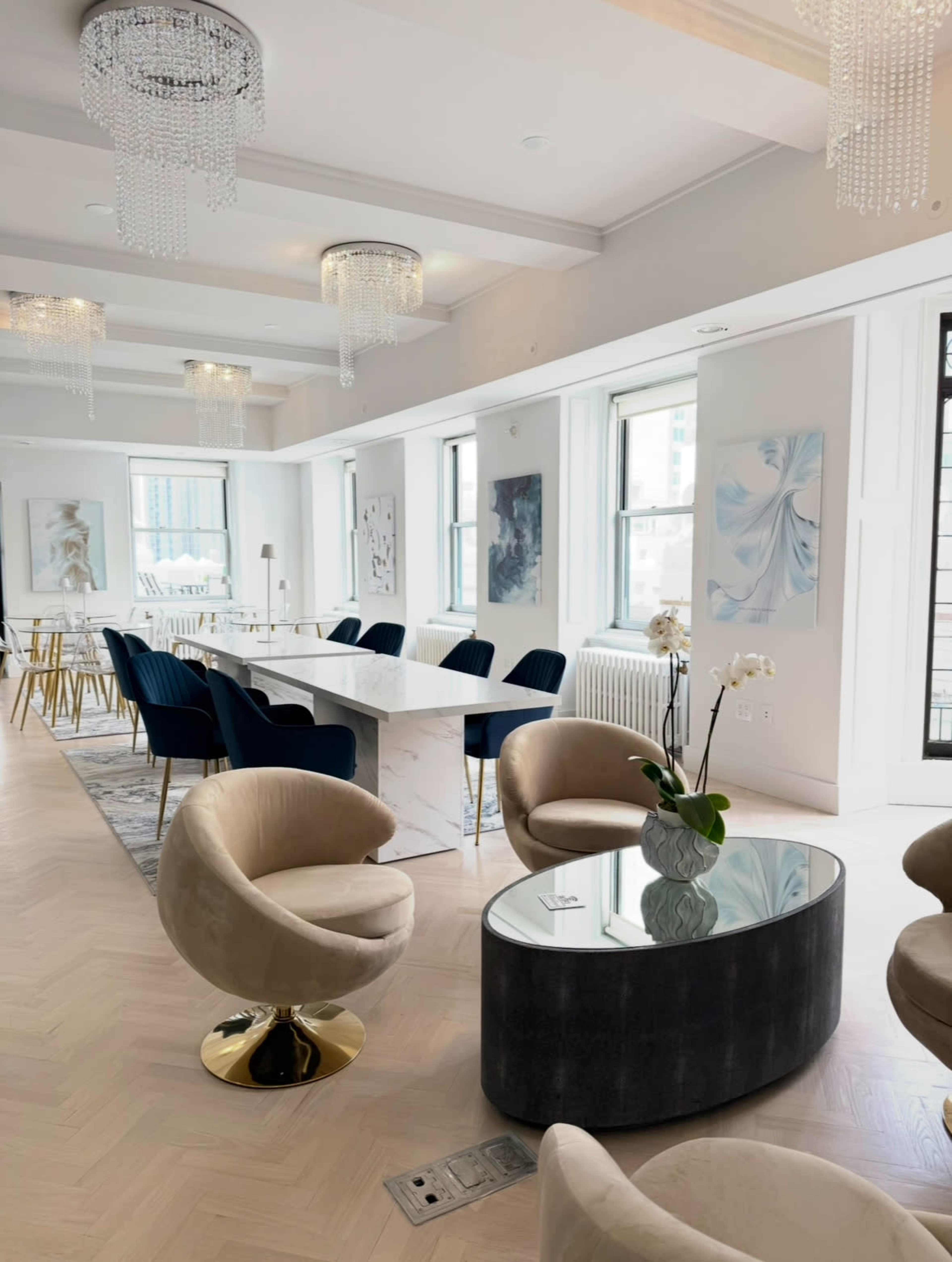 The image shows a modern, elegantly designed lounge area with round chairs, a coffee table, and a dining space featuring a long marble table and blue upholstered chairs.
