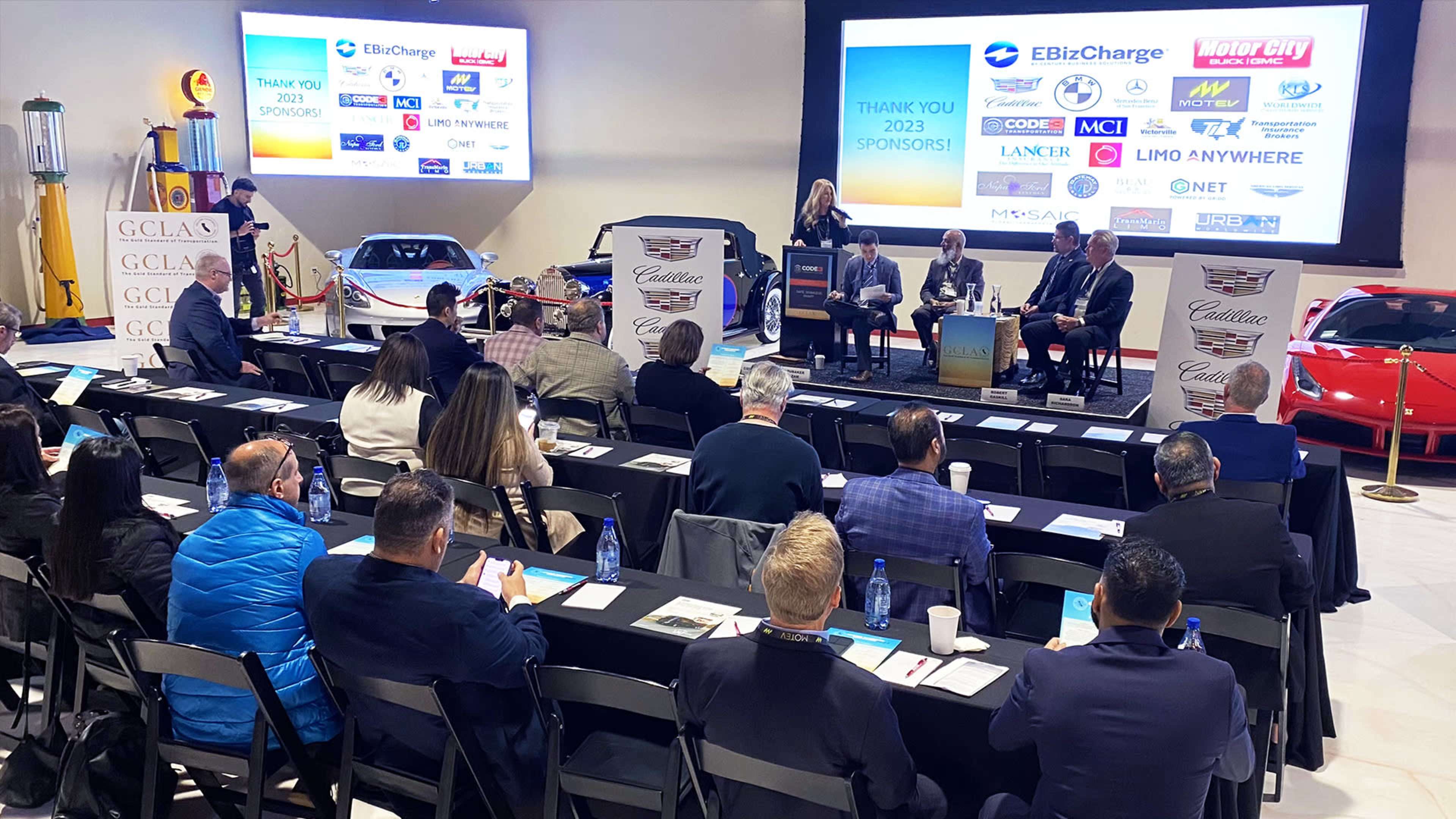 A panel discussion is taking place in front of an audience at an event featuring several classic cars and promotional screens in the background.