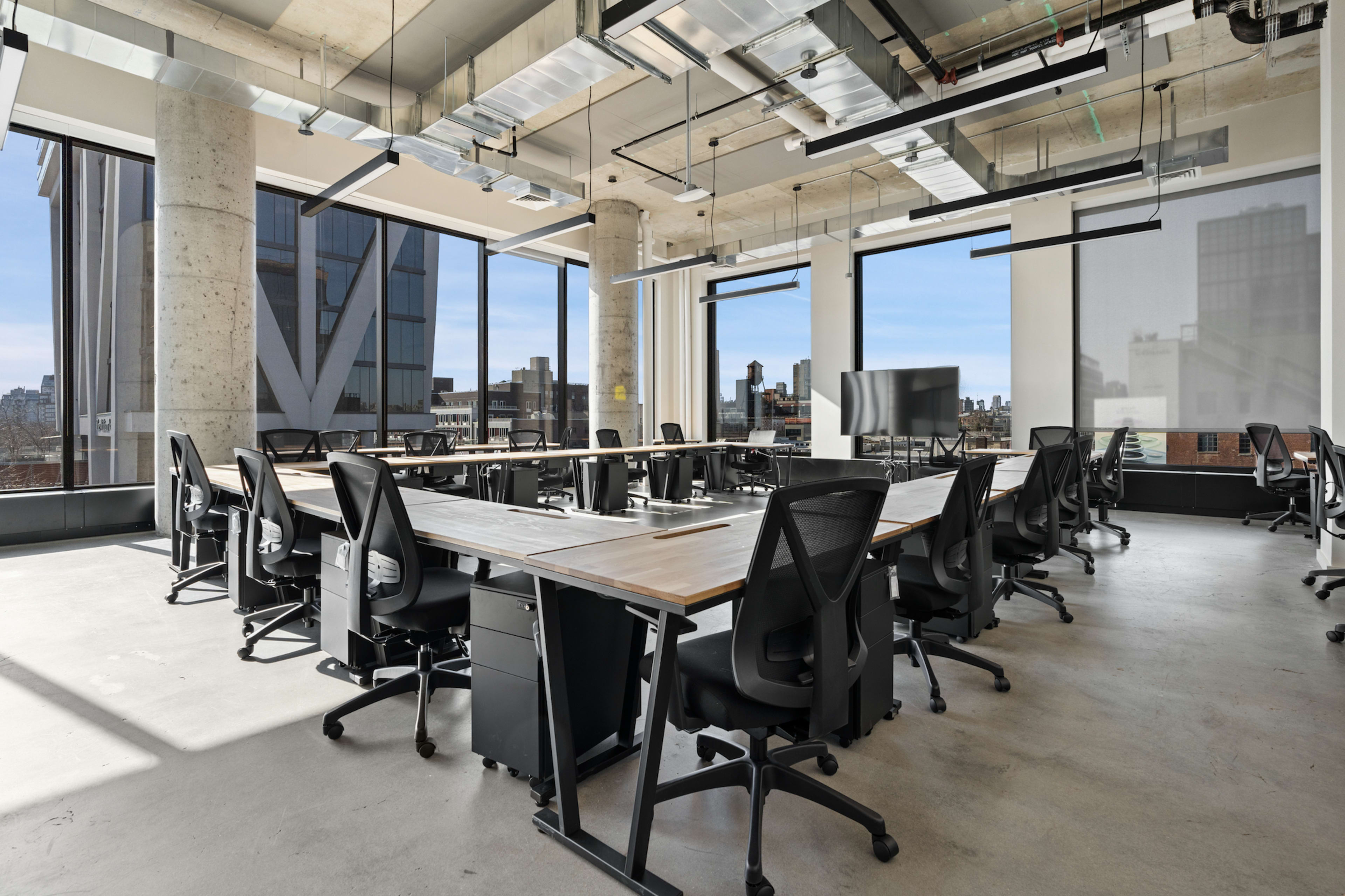 The image shows a modern office space with multiple long tables and ergonomic chairs arranged around them, featuring large windows that provide a view of a city skyline.
